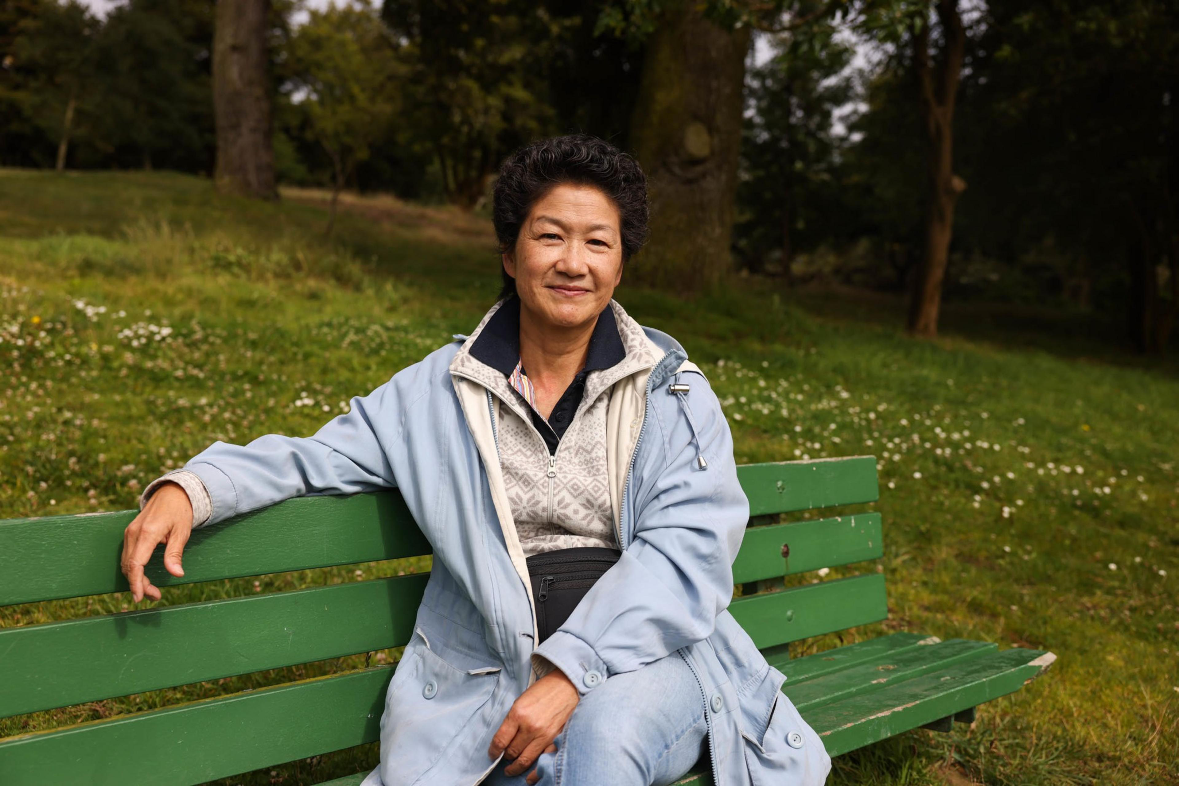 A woman wearing a light blue jacket sits on a green bench in a park, smiling gently. Behind her are green trees and a grassy area with scattered white flowers.