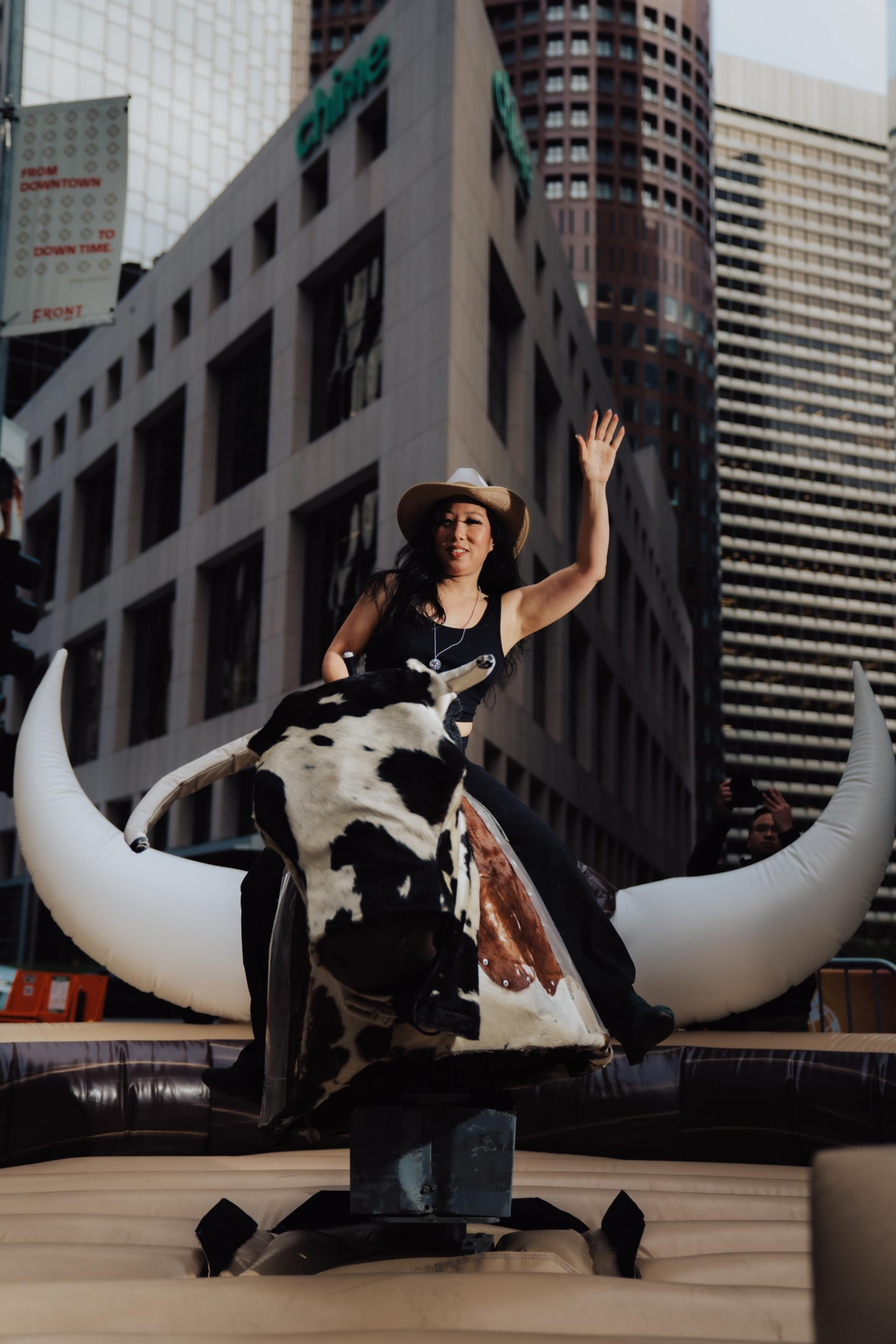 A person wearing a cowboy hat rides a mechanical bull in an urban setting, surrounded by tall buildings. They appear to be waving with one hand.