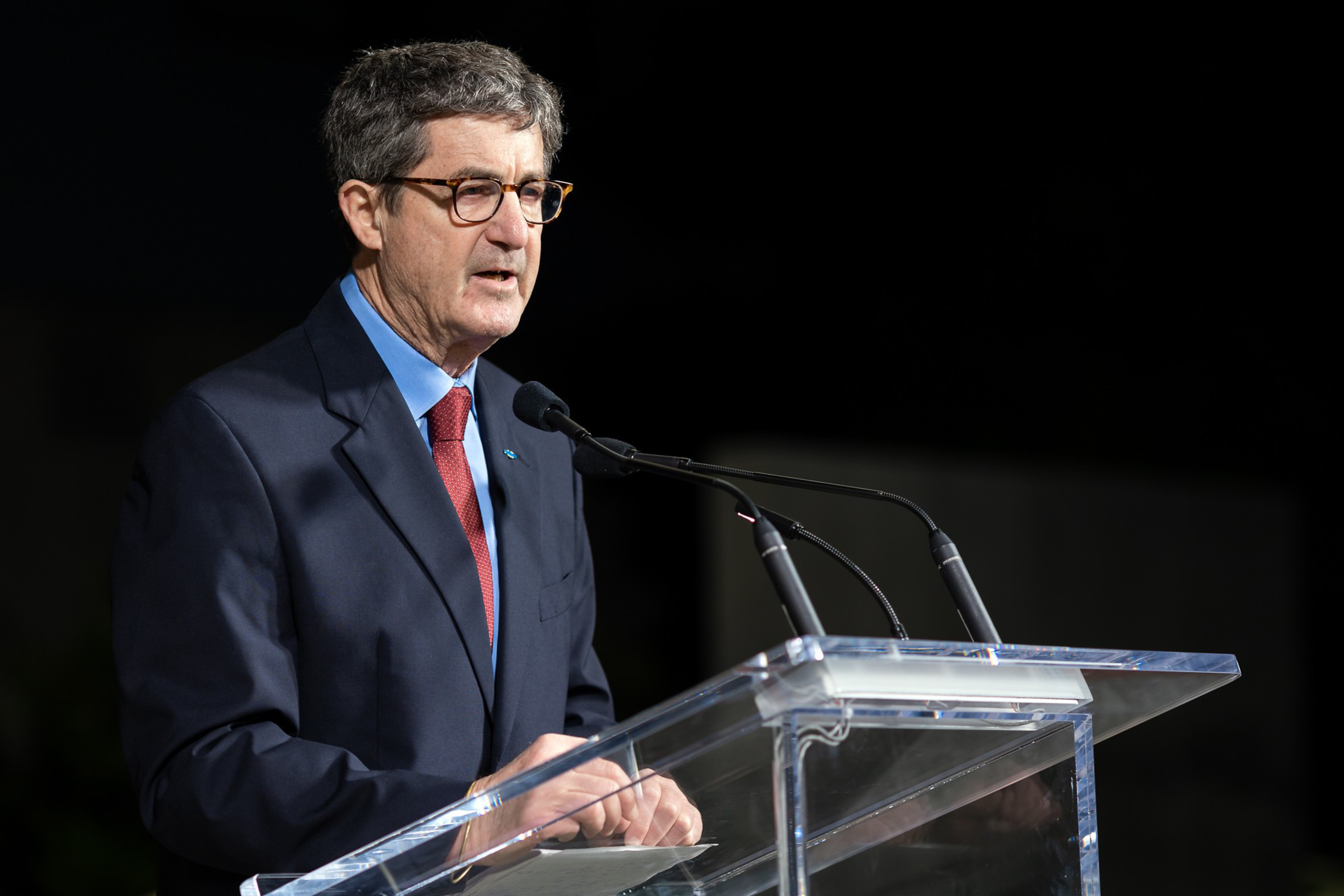 A man in a suit and glasses speaks at a transparent podium with microphones.