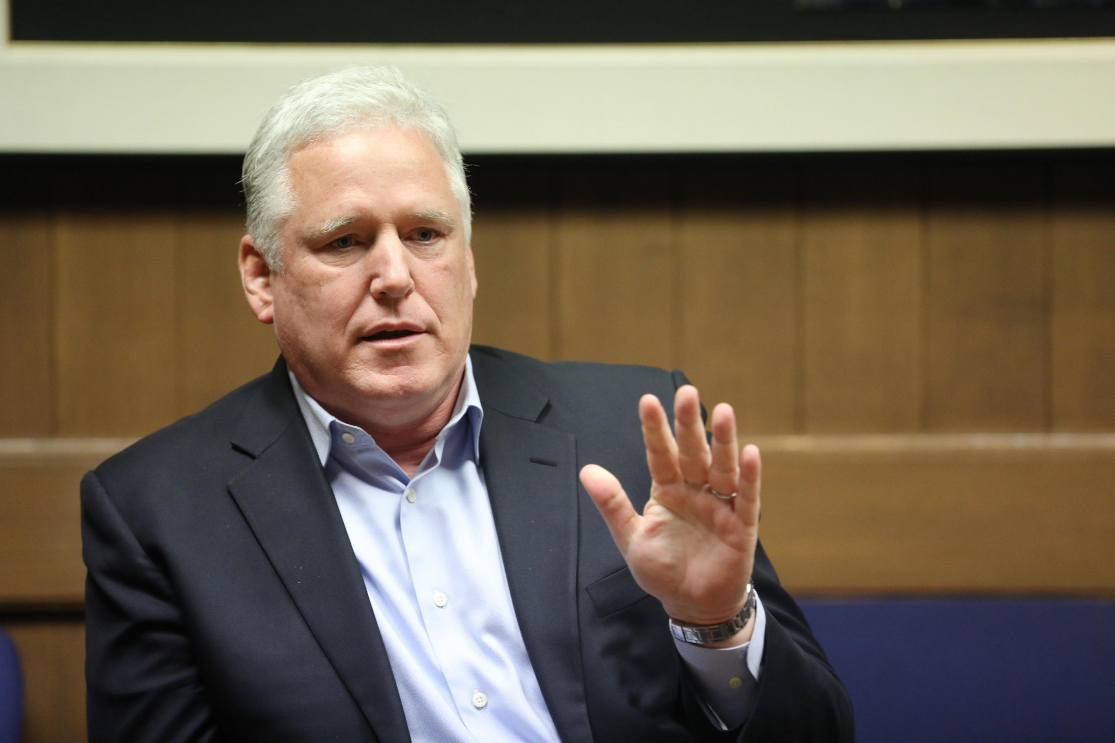 A man, John Dennis, raises his right hand slightly gesticulating while sitting in a board room.