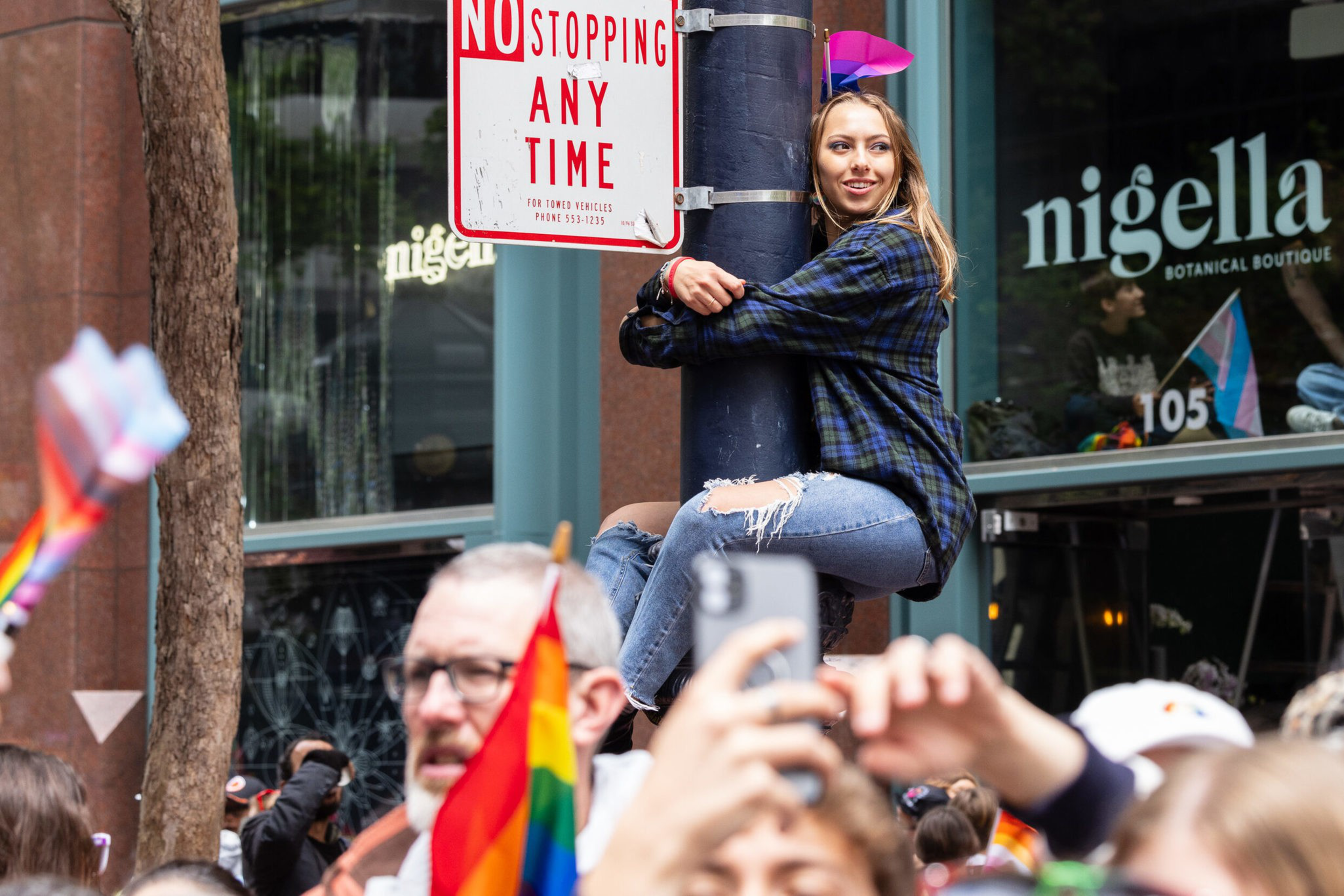 A young woman wearing a purple and blue plaid shirt and ripped jeans clings to a black pole beneath a "No Stopping Any Time" sign, smiling amid a crowd with pride flags.
