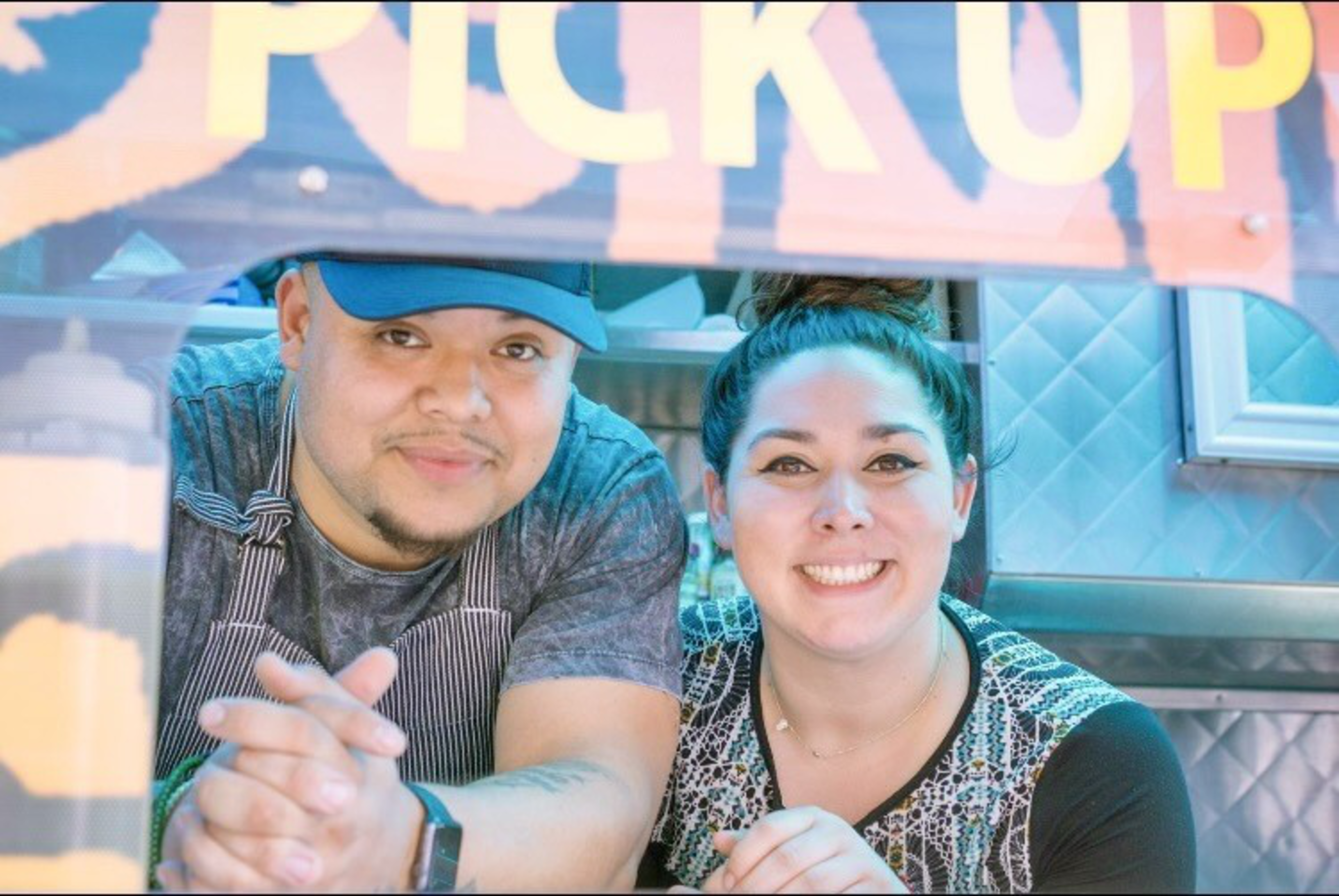 A man and woman smile warmly behind a window labeled "PICK UP" inside a brightly lit food truck or kitchen setting.