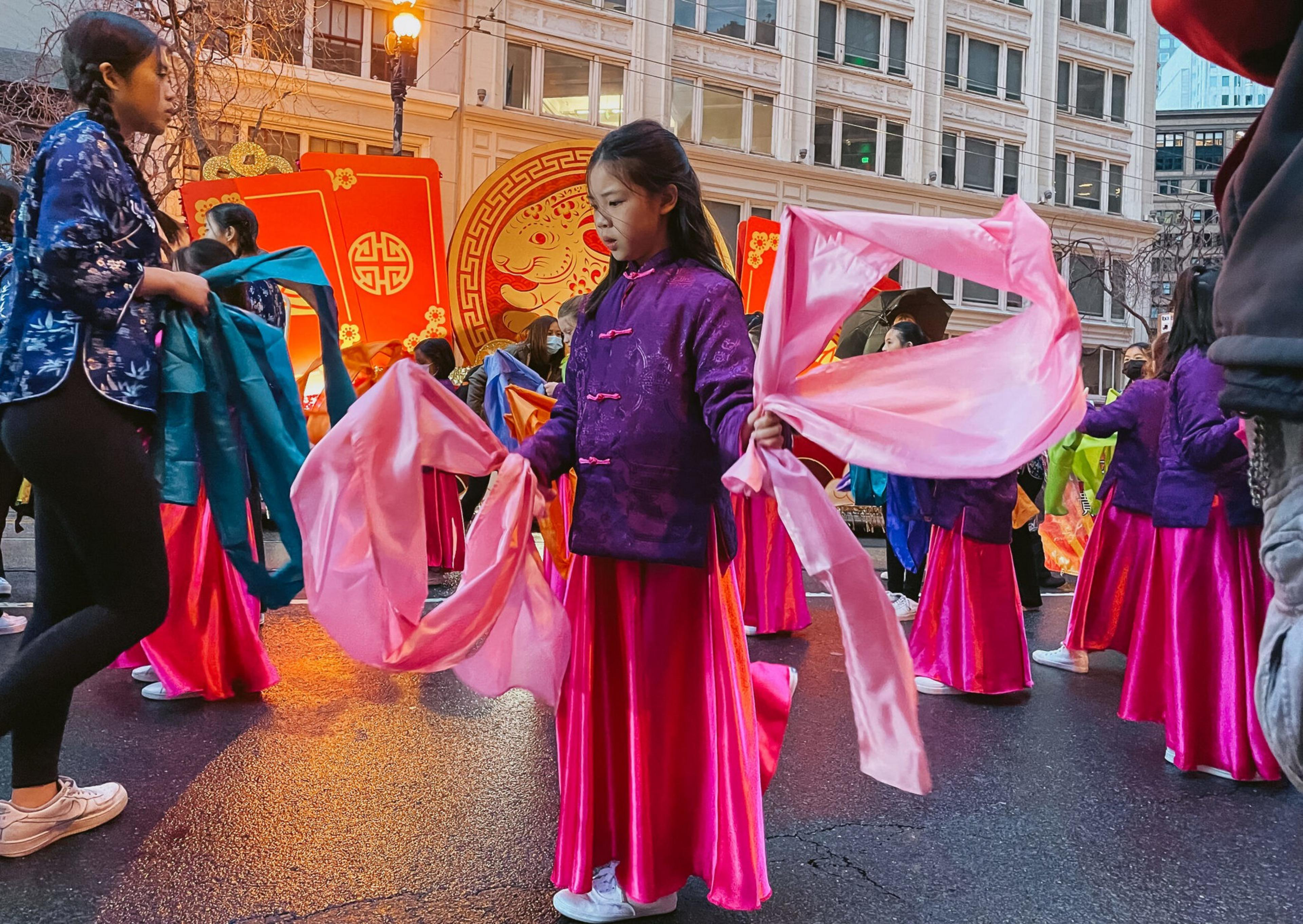 Young girls in colorful traditional attire perform a dance with flowing fabric ribbons on a city street during a festive event.
