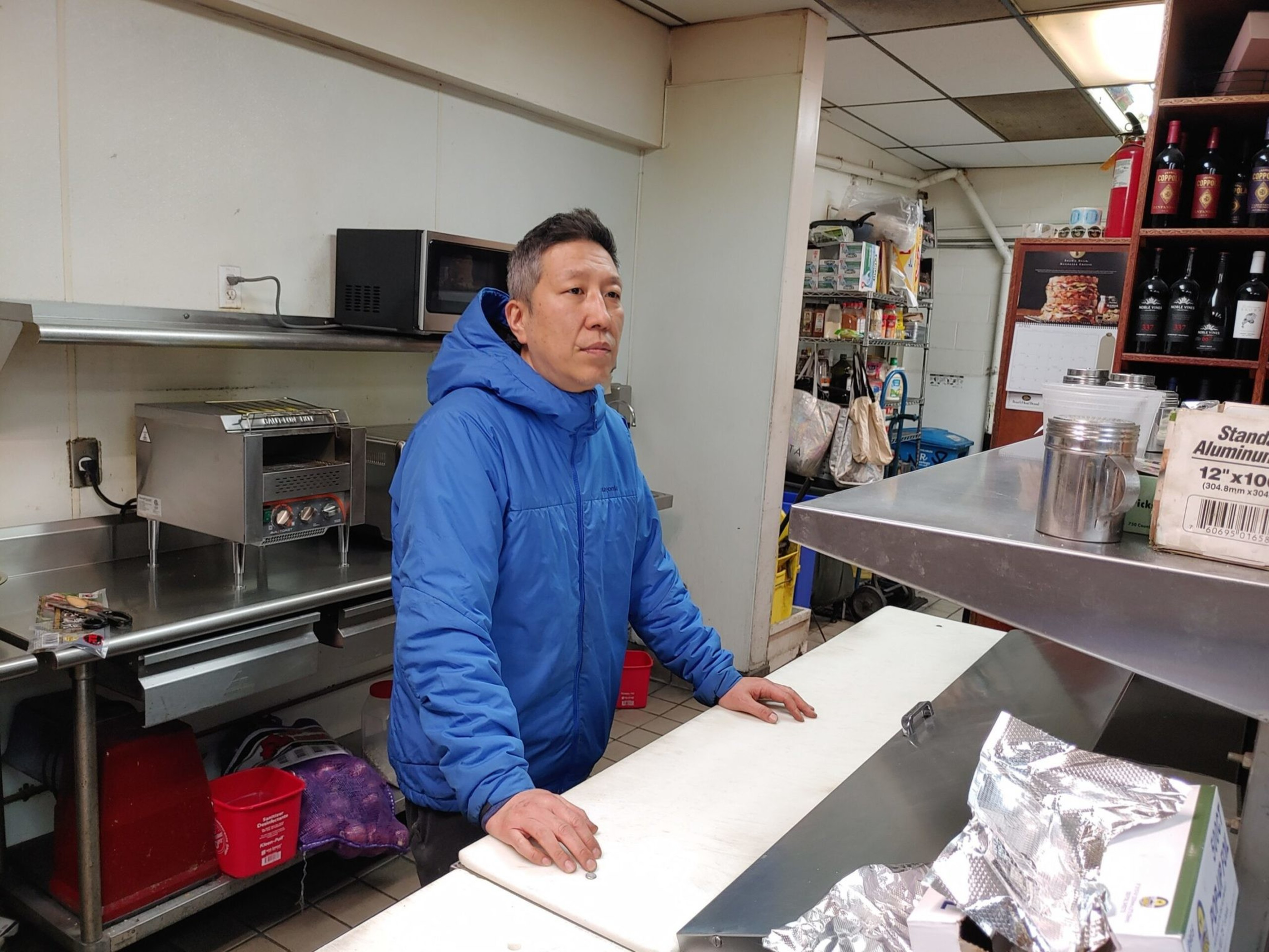 A man in a blue jacket stands behind a counter in a kitchen or food prep area, surrounded by appliances, food supplies, and shelves with bottles.
