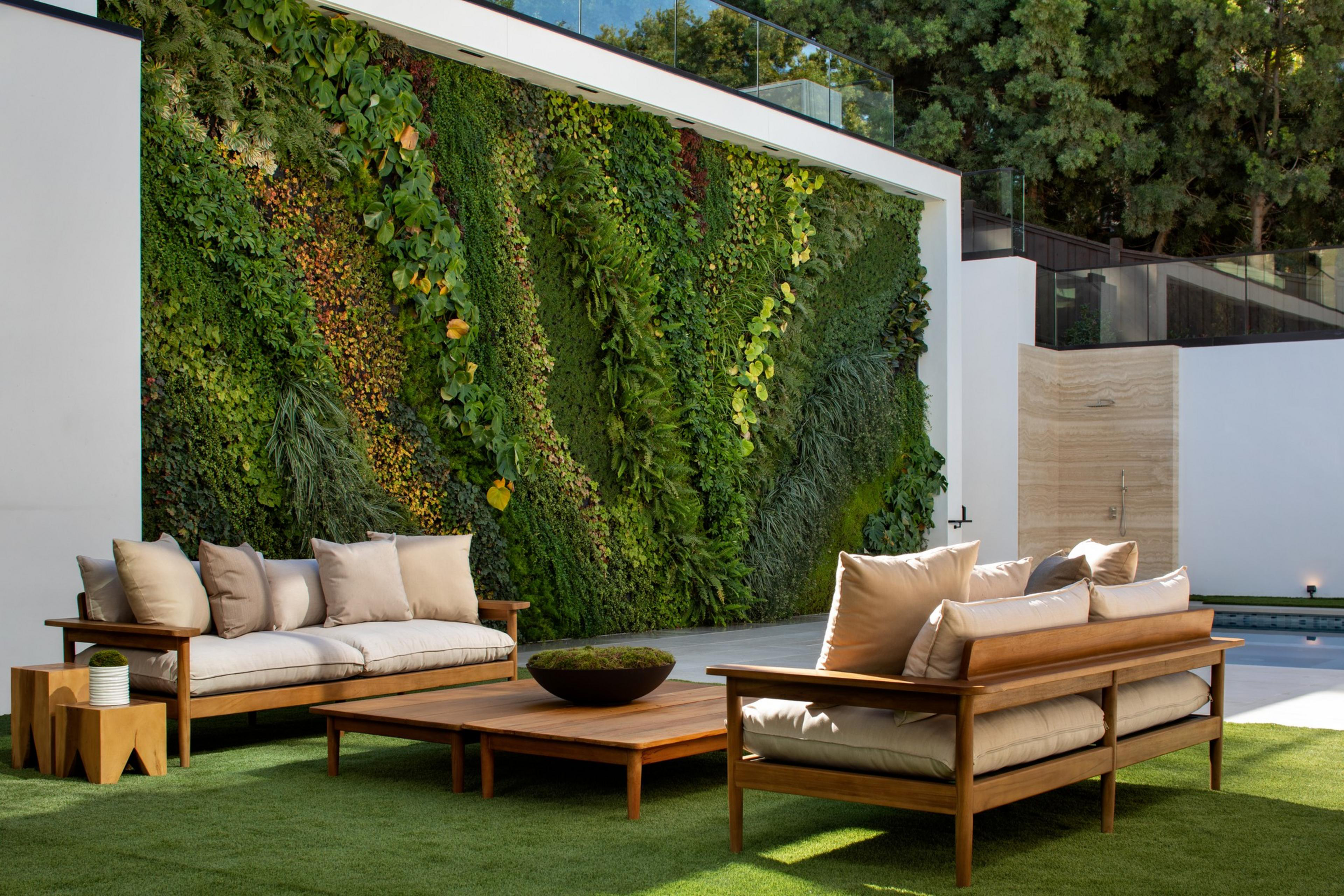 Two wooden couches with beige cushions surround a low wooden table on artificial grass, set against a tall, lush green vertical garden wall.