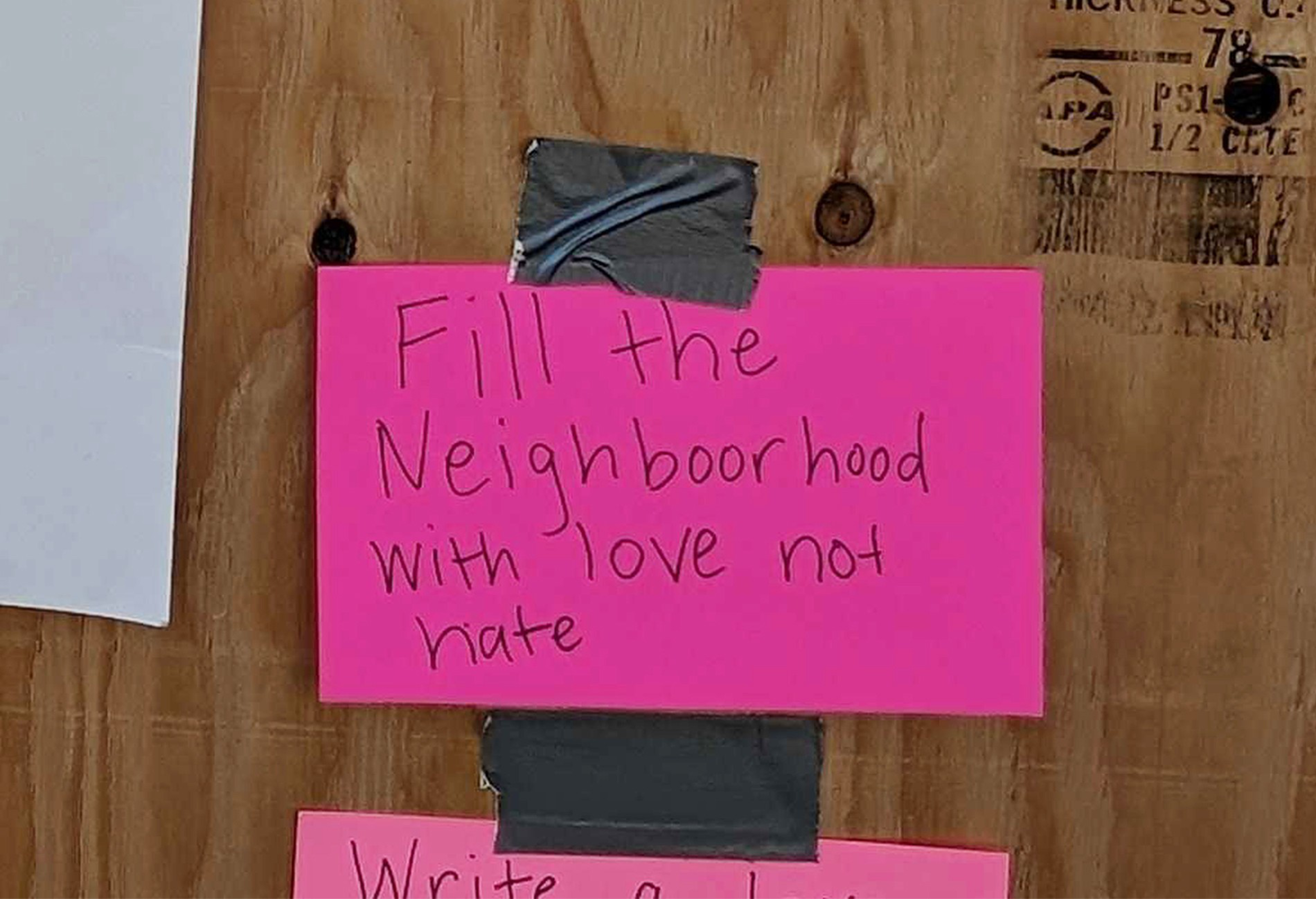A neon pink card taped to a wooden board reads, &quot;Fill the Neighboorhood with love not hate,&quot; in handwritten text.