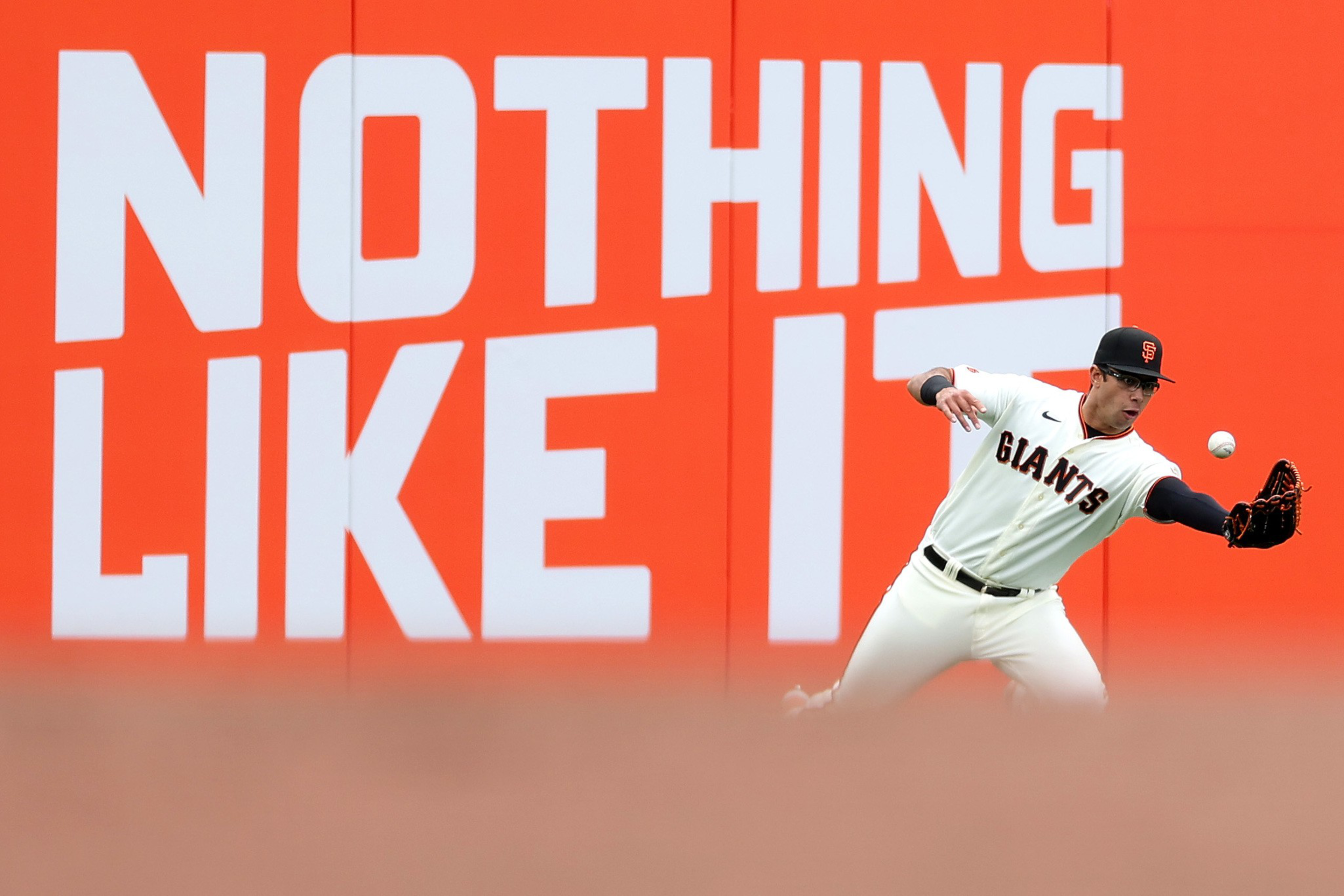 A baseball player in a Giants uniform reaches out with a glove to catch a ball near a bright orange wall with large white letters spelling "NOTHING LIKE IT."