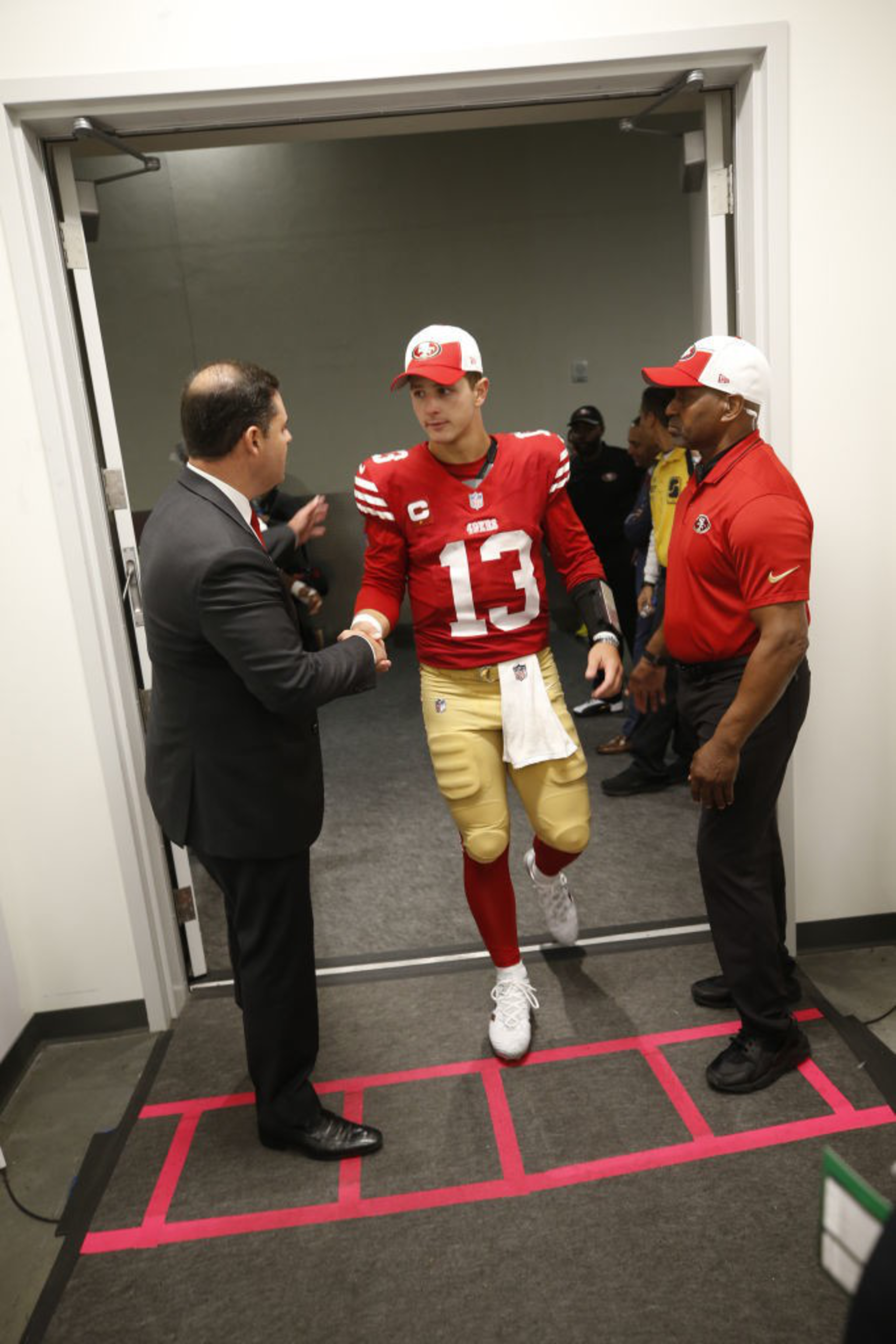 A football player in a red and gold uniform shakes hands with a man in a suit. Another man in a red shirt stands nearby. They are in a hallway.