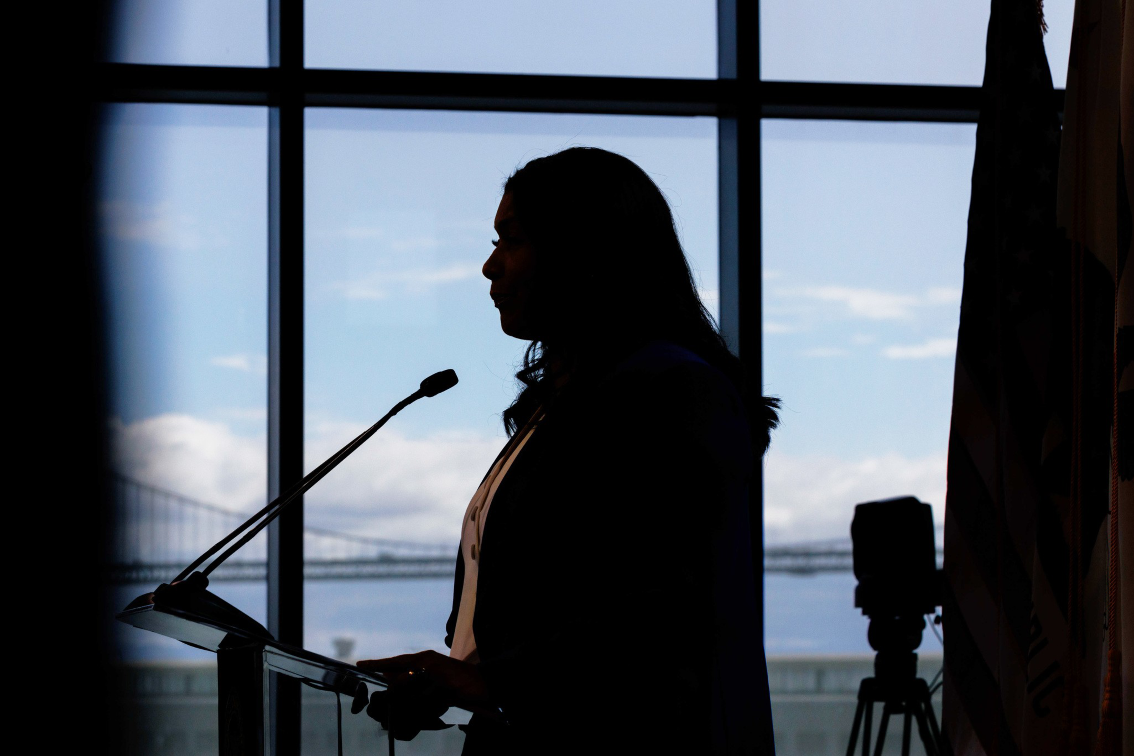 A silhouetted person stands at a podium with a microphone. The background shows a large window with a bridge and a clear sky, while a camera is visible to the side.