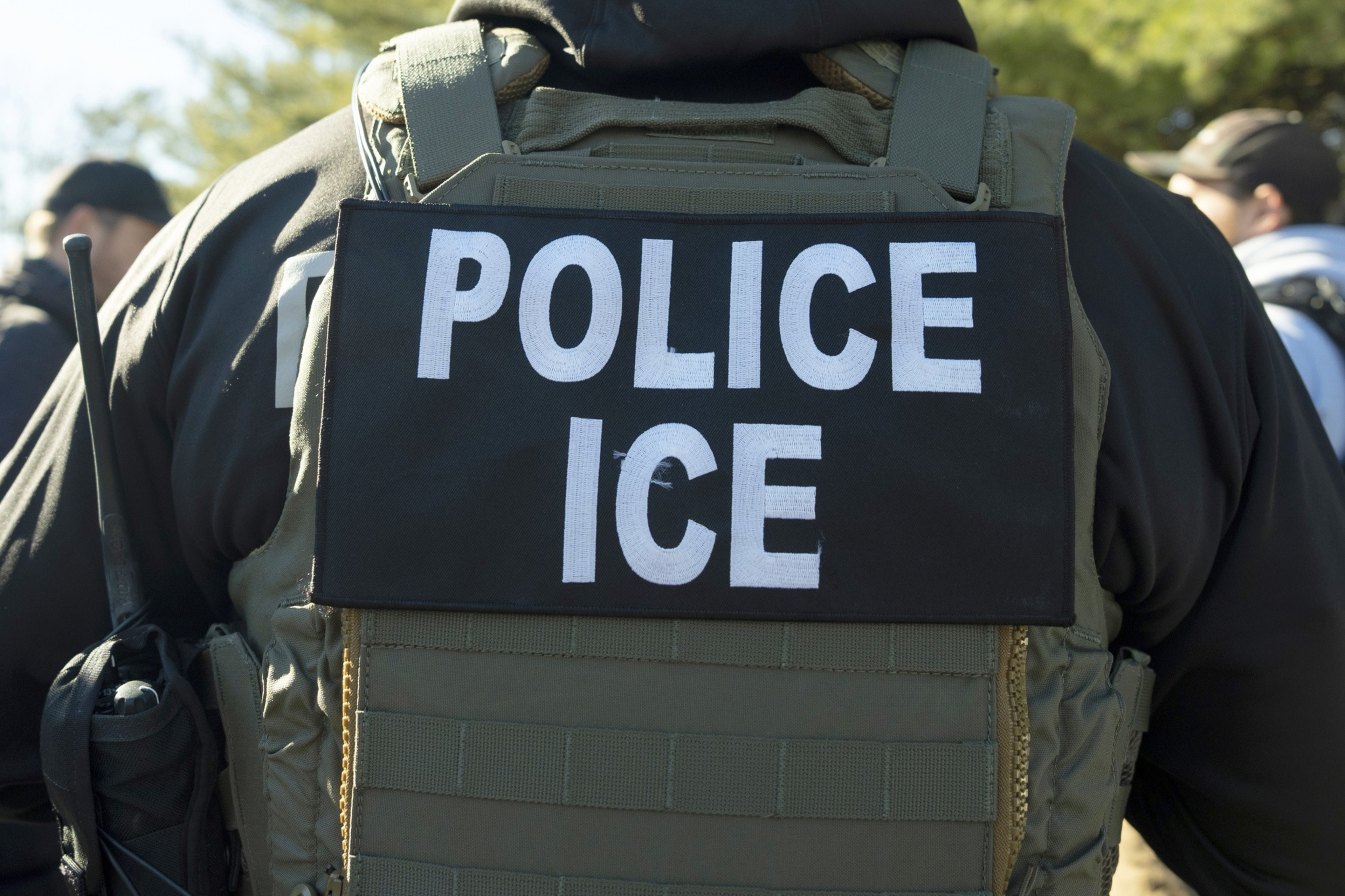 A person is wearing a tactical vest with a patch labeled "POLICE ICE." The vest is dark, and there are others in the background, possibly outdoors.