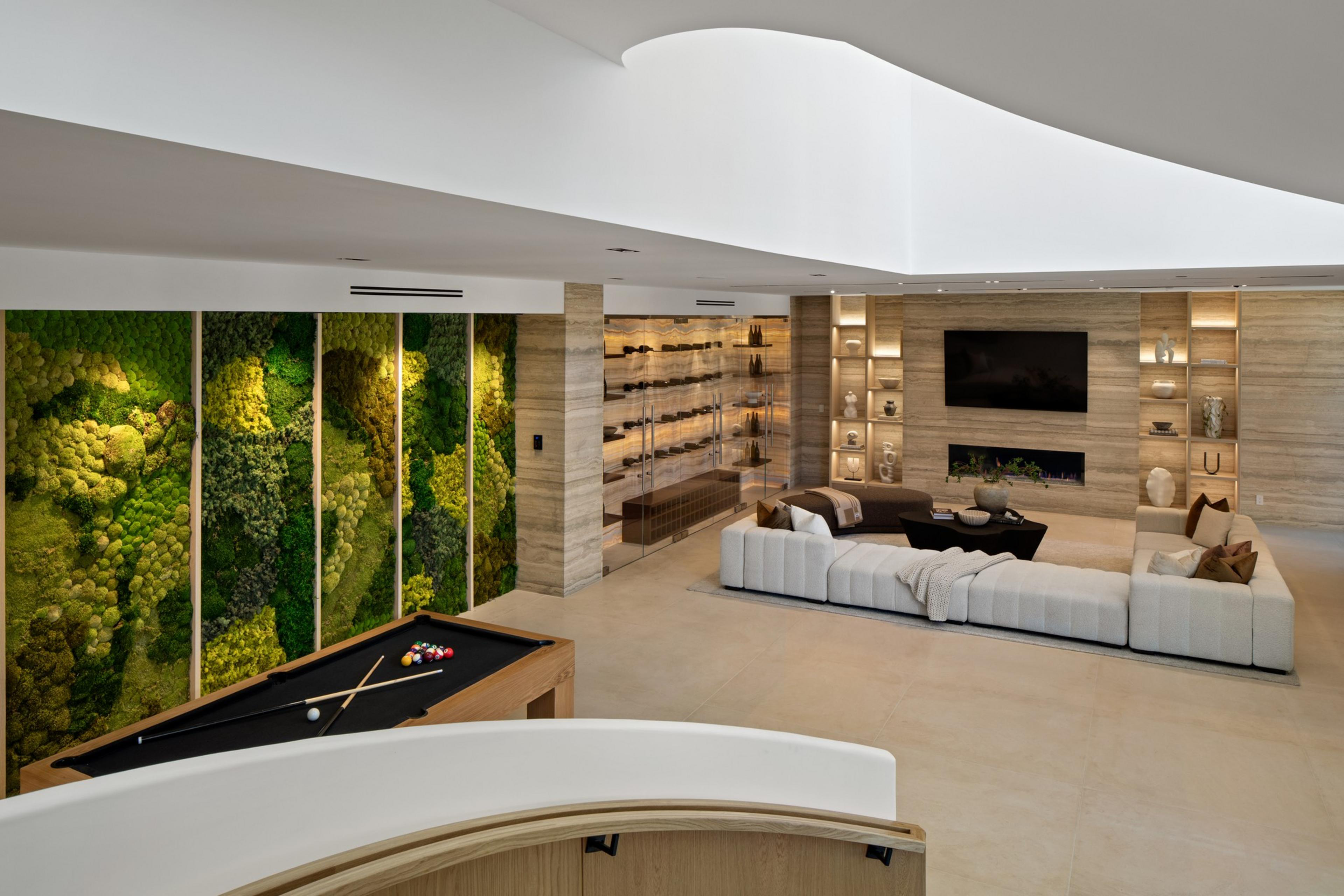 A modern living space features a white sectional sofa, a black pool table, a moss wall with vertical panels, and a media wall with a TV and fireplace.
