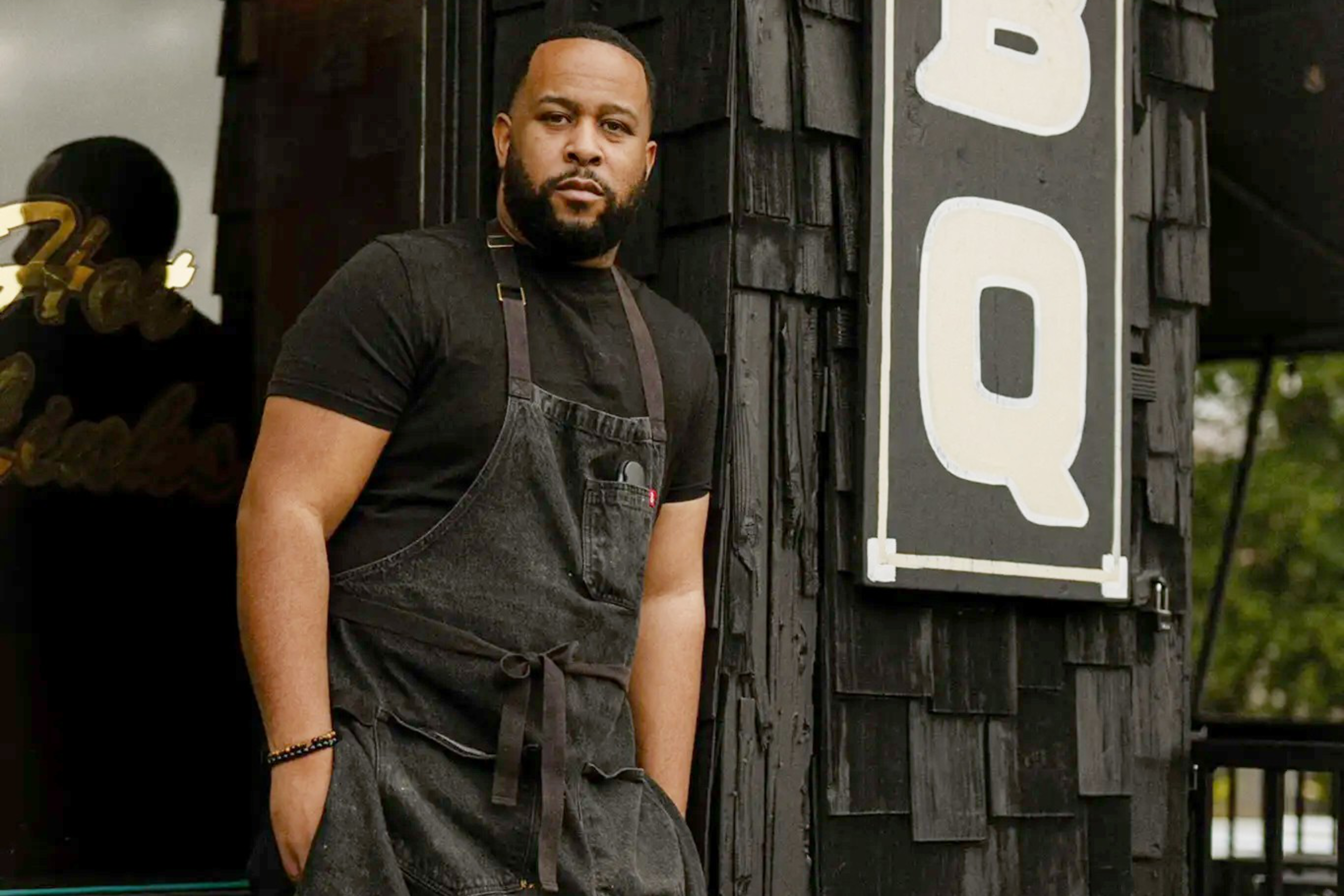A man in a black apron stands confidently outside a BBQ restaurant. He's leaning against a building with black wood siding and a large "BBQ" sign.