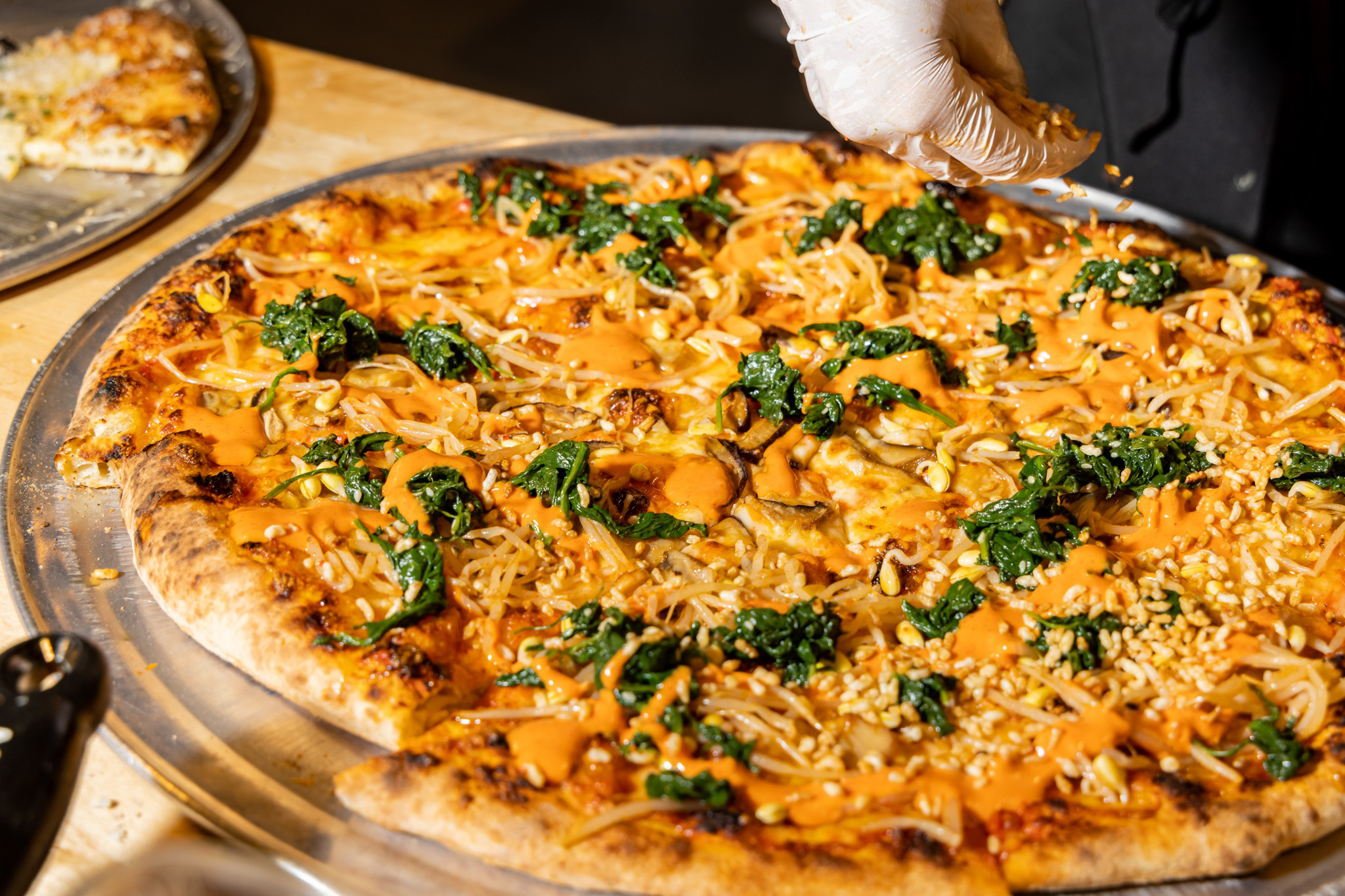 A pizza topped with spinach, bean sprouts, orange sauce, and sprinkled sesame seeds, being garnished by a gloved hand.