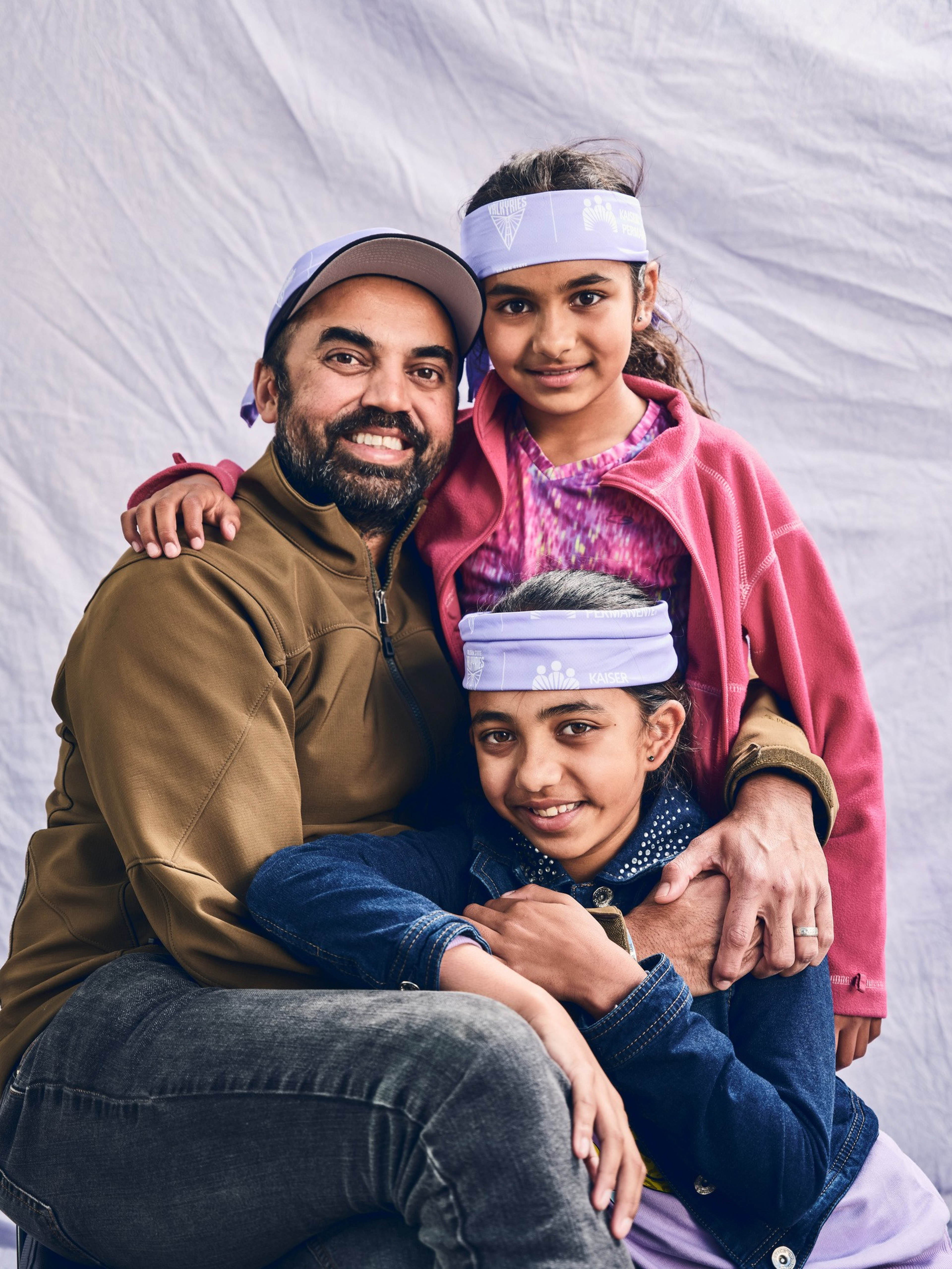 A man and two girls smile warmly, wearing casual clothes and matching light purple headbands, with the girls embracing the man affectionately.