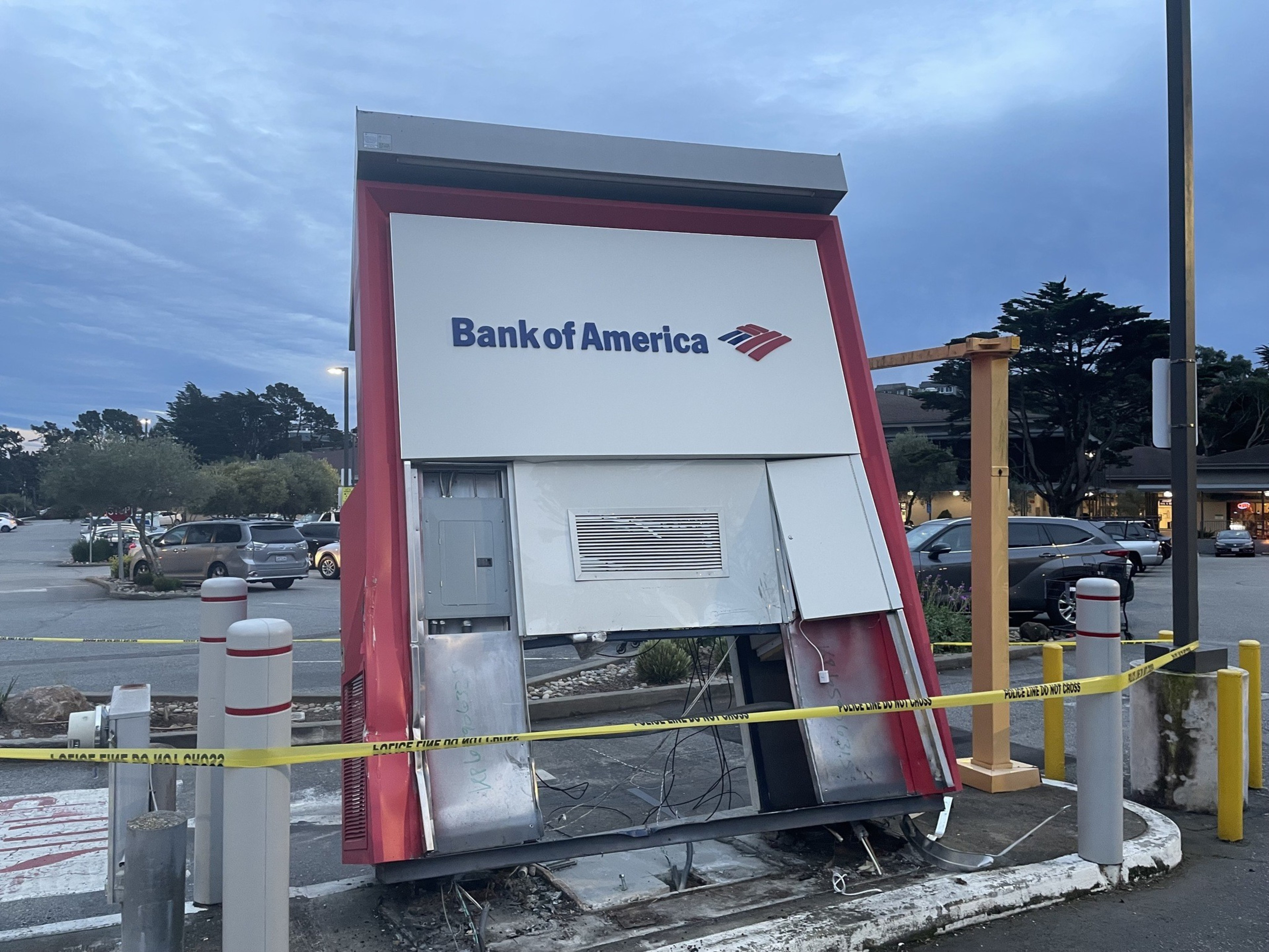 A damaged Bank of America ATM is cordoned off with yellow caution tape in a parking lot during twilight.