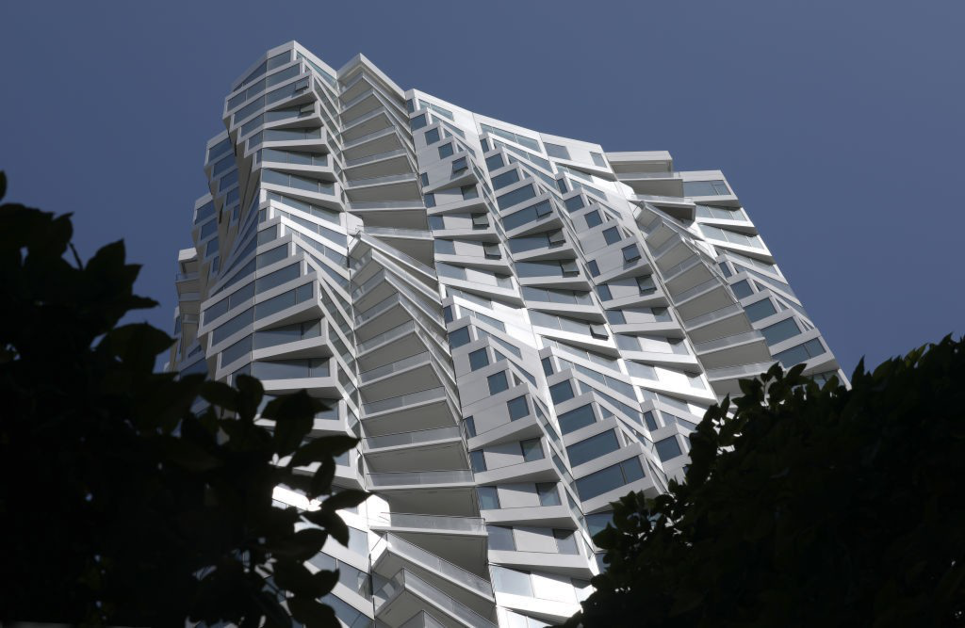A modern, angular high-rise building with reflective glass panels and twisted geometric shapes rises against a clear blue sky, framed by dark tree silhouettes.