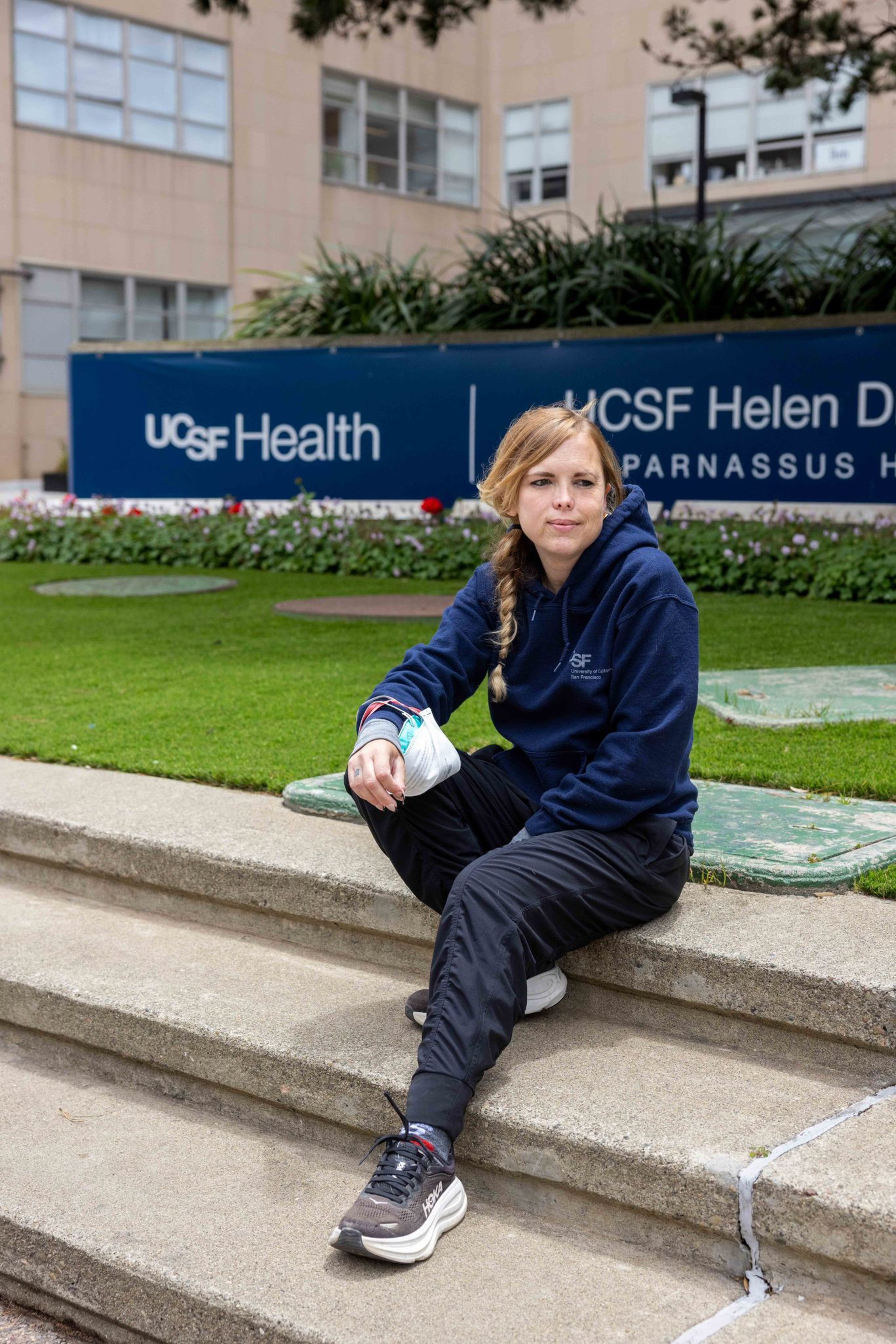 A woman in a navy hoodie and black pants sits on concrete steps holding a face mask, with a UCSF Health hospital sign and plants behind her.