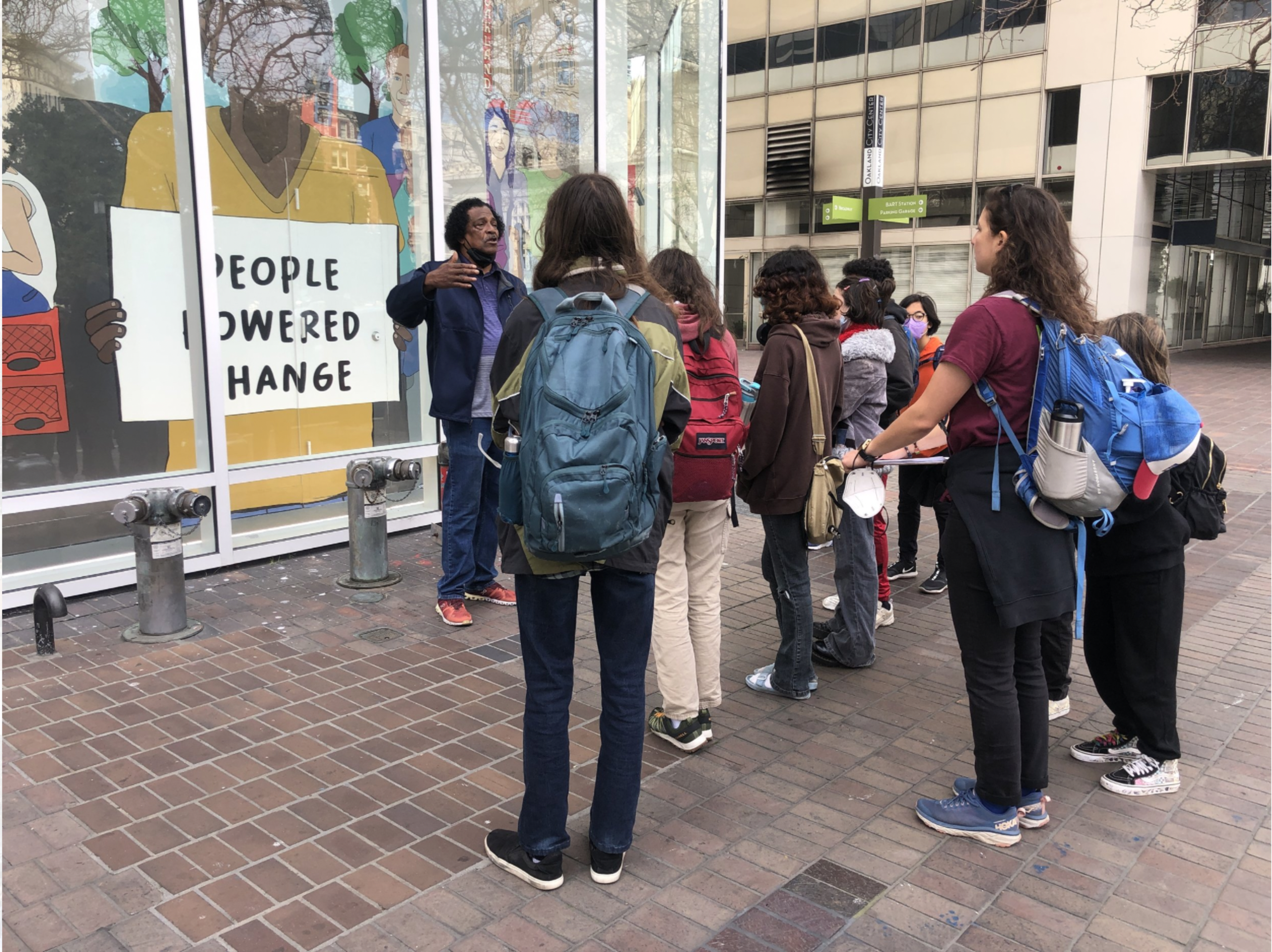 A group of people with backpacks stand on a brick sidewalk listening to a man speaking near a window mural reading "People Powered Change."
