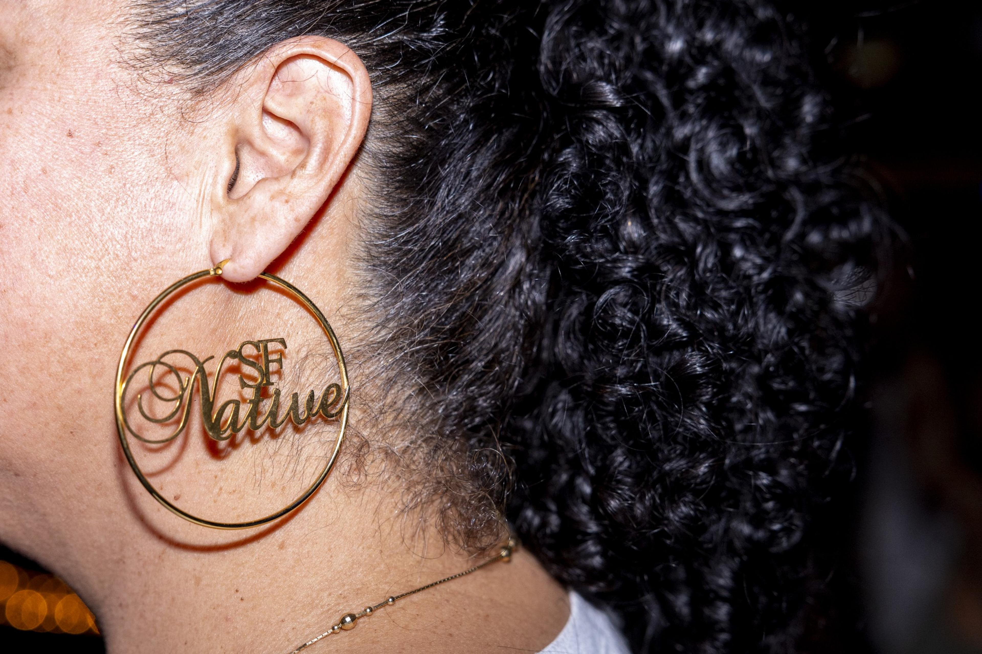 The image shows a close-up of a person's ear with a large, gold hoop earring that reads &quot;SF Native&quot; in a cursive font. The person has curly dark hair.