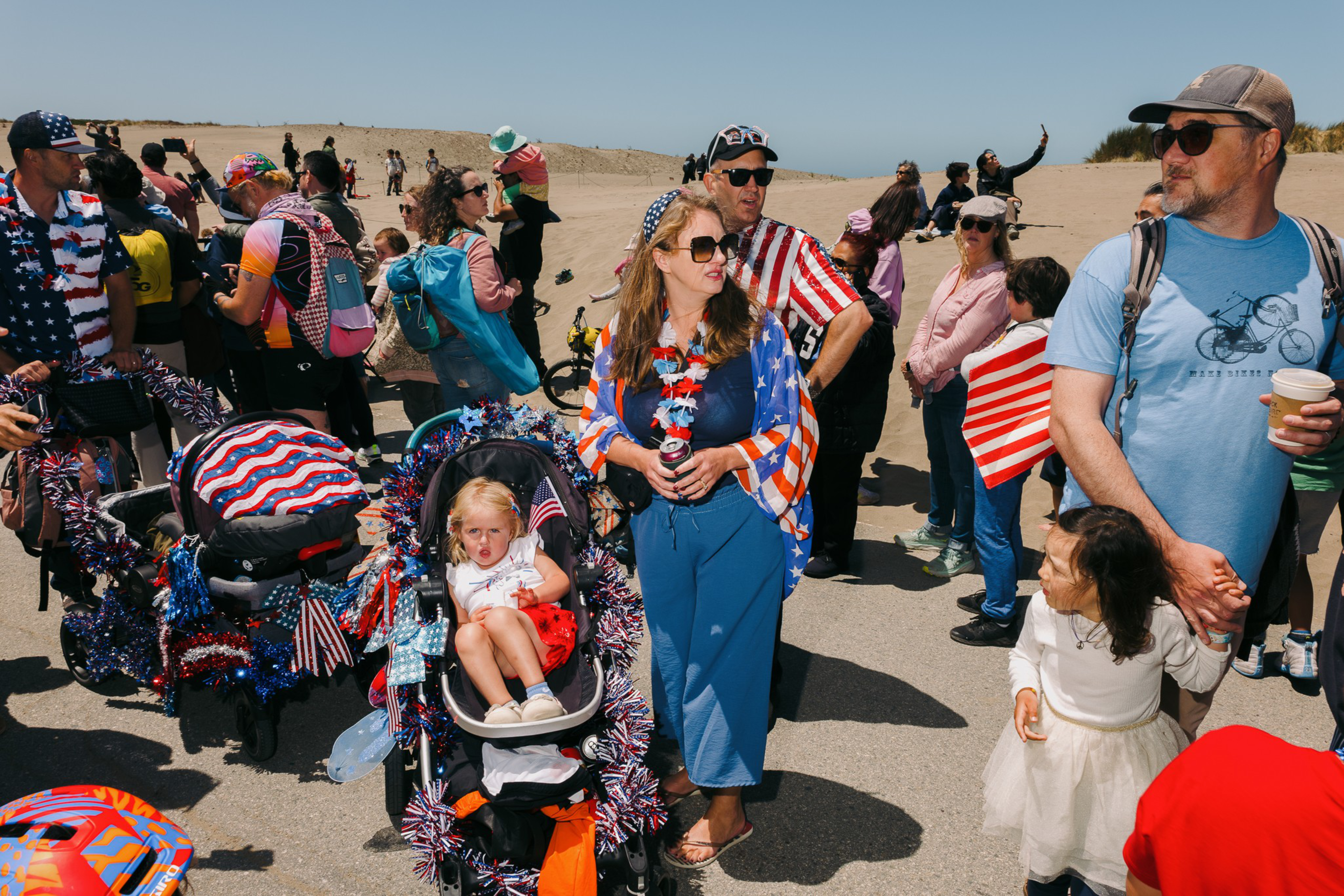 The image shows a crowd at a beach, with people wearing American flag-themed outfits. There are children in decorated strollers and some holding flags. It's a sunny day.