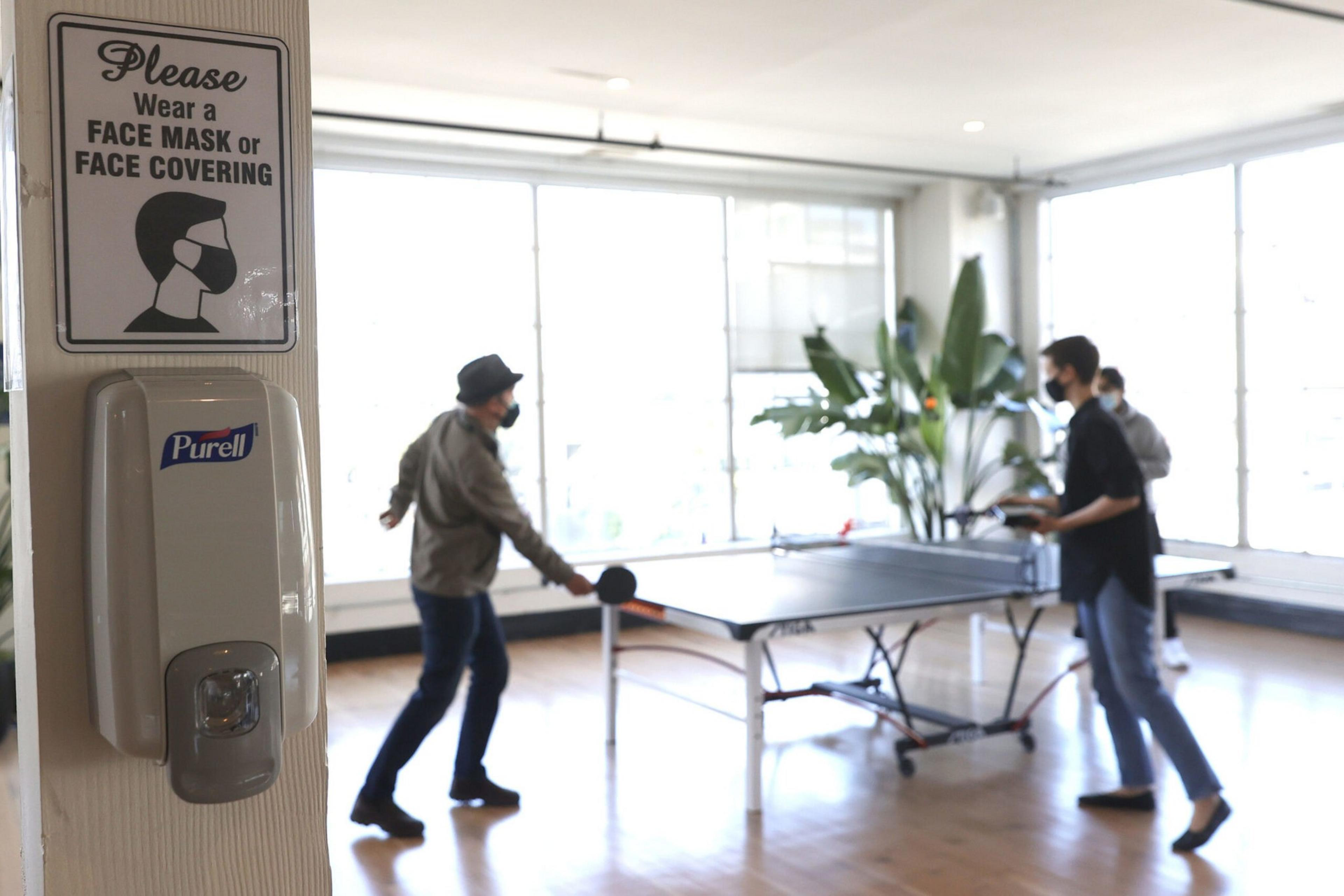 A hand sanitizer dispenser and a mask-wearing sign are in the foreground, while three masked people play table tennis in a bright room.