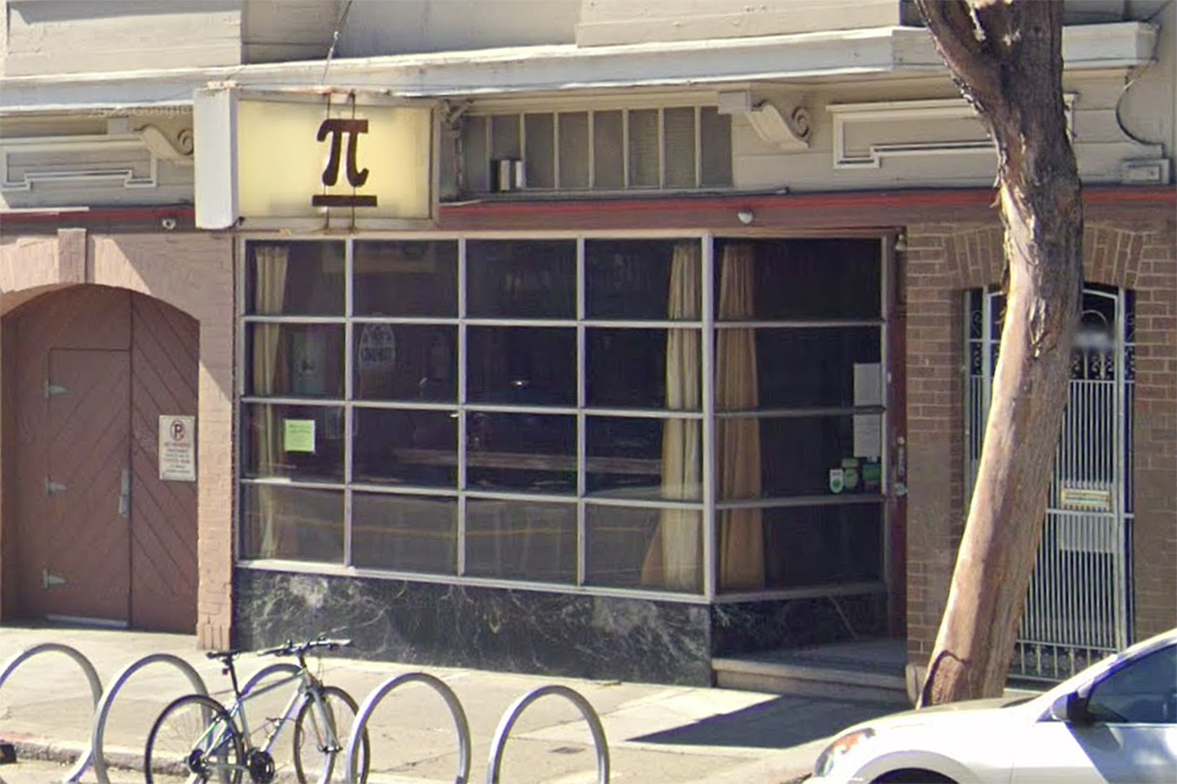 A building with a large windowed storefront featuring beige curtains, a bike rack with a bike in front, and a sign above displaying the pi symbol (π).