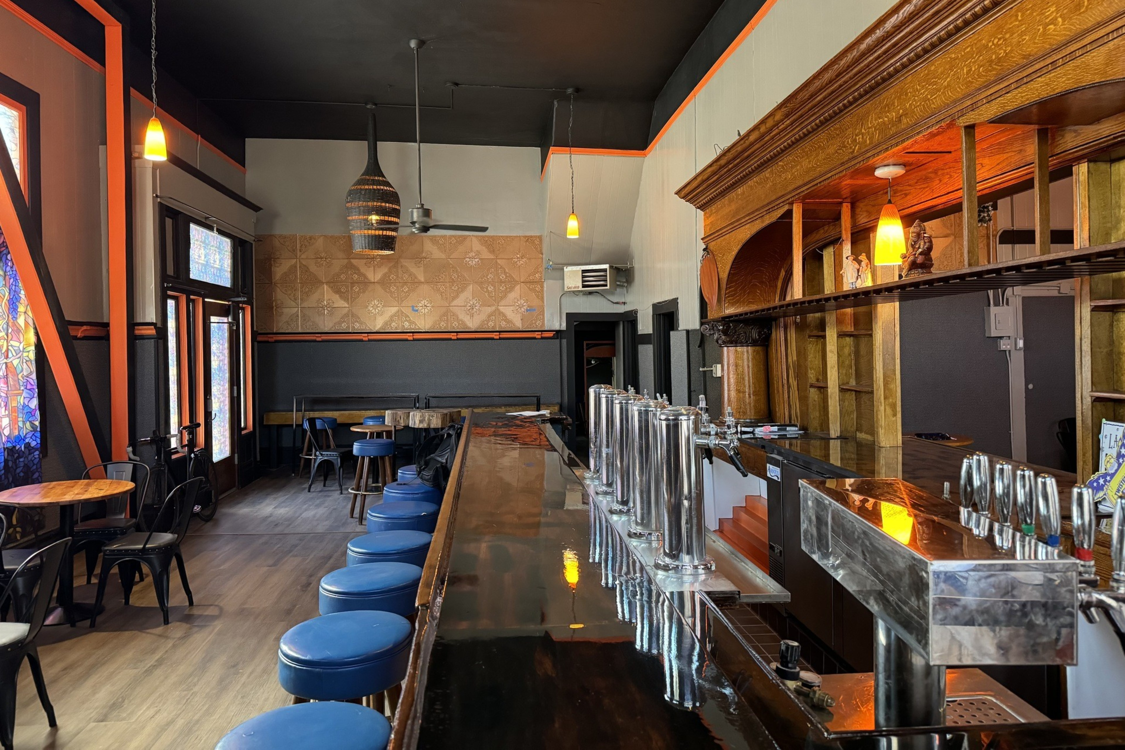 The image shows a bar with wooden decor, orange accents, pendant lights, a mirrored counter, and blue stools. Stained glass windows and mosaic tiled walls adorn the space.