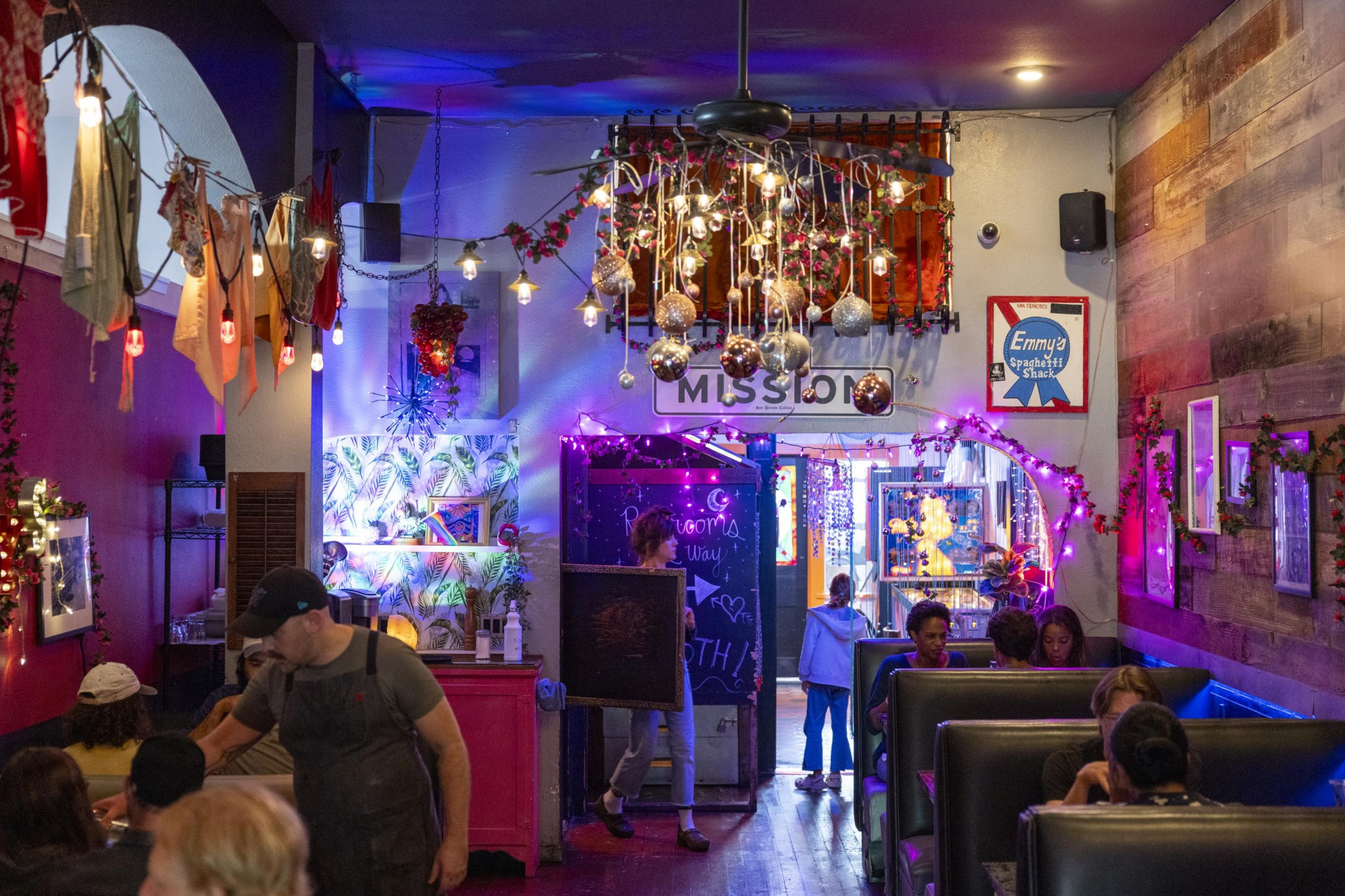 The image shows a cozy, eclectic restaurant with vibrant decorations, string lights, and hanging ornaments. People are seated in booths, and a server is attending to a table.