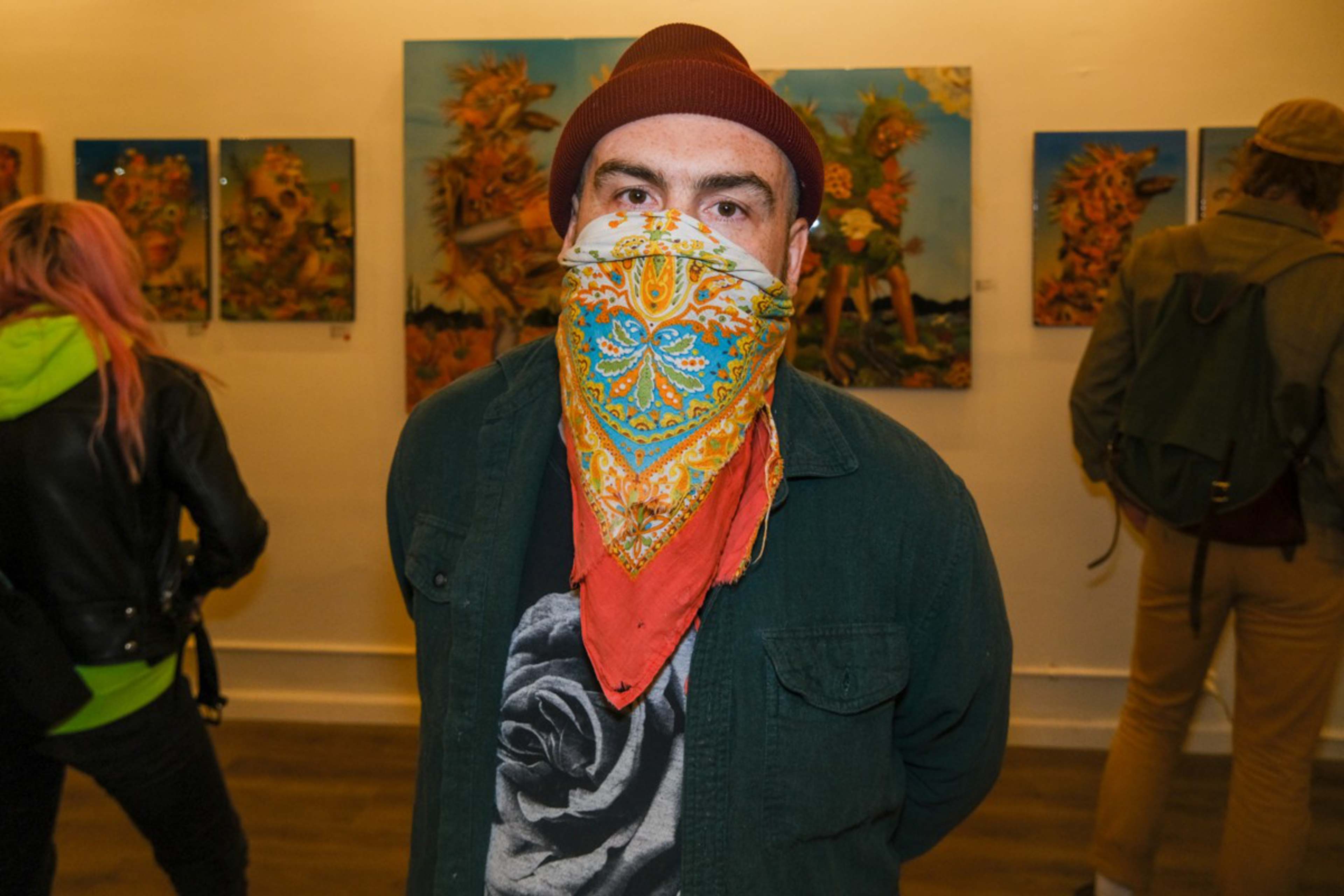 A man wearing a maroon beanie and a colorful bandana over his face stands in an art gallery with paintings and two other people in the background.