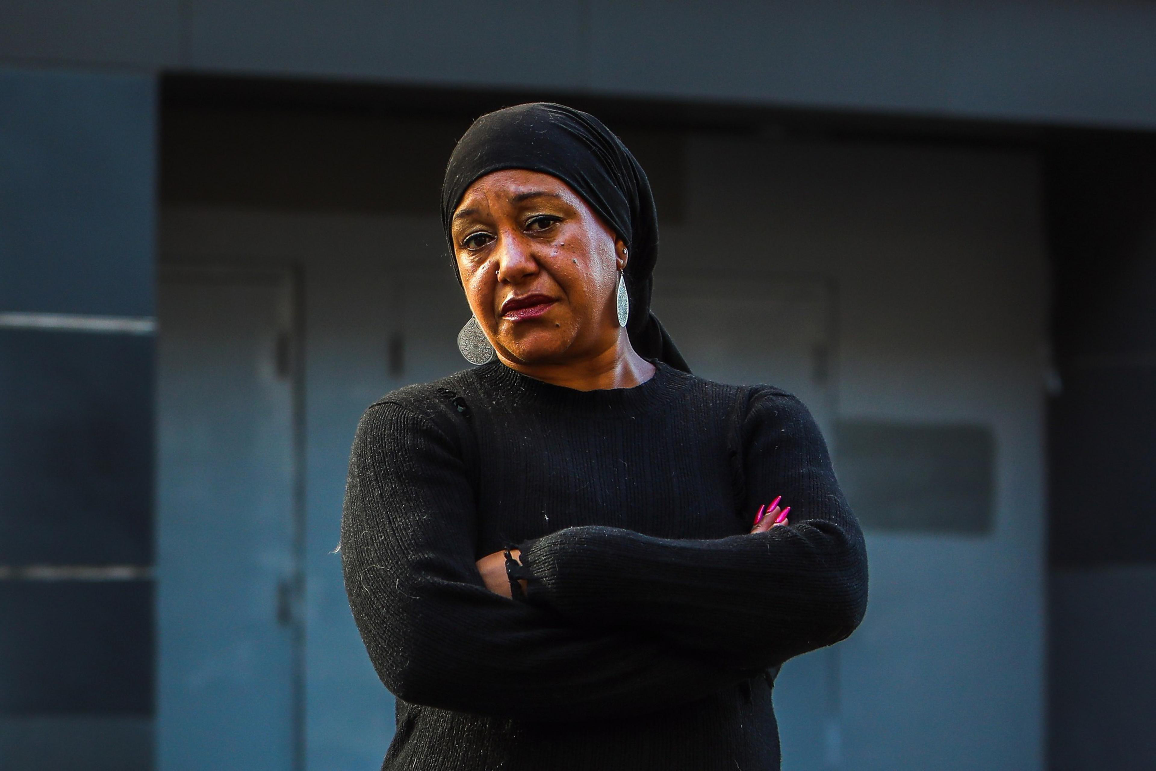 A woman wearing a black headscarf and sweater stands with arms crossed, looking serious against a blurry gray background.