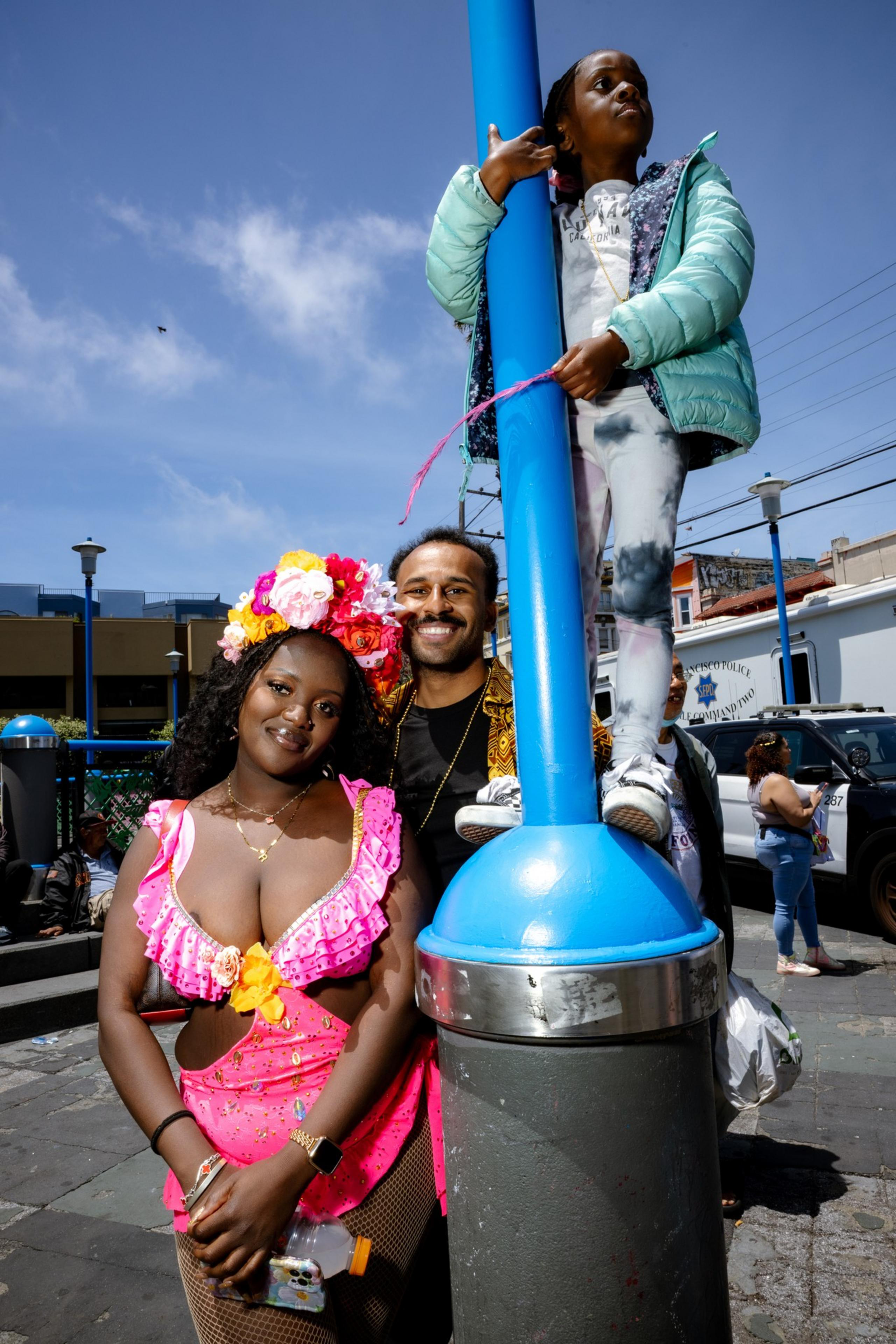 A woman in a vibrant pink outfit smiles with a man and child by a blue pole. The background shows an urban setting under a clear blue sky.
