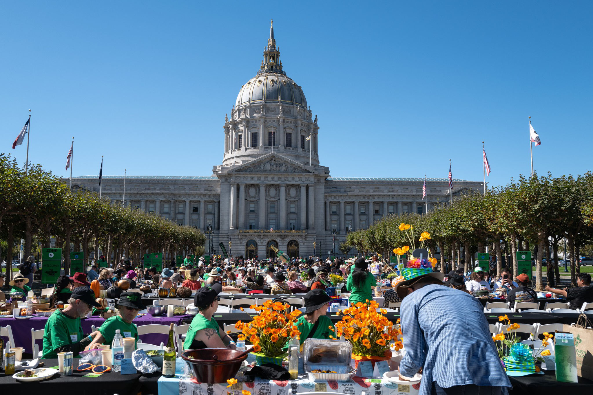 Hundreds of people gather at long tables with orange flowers in front of a grand domed government building on a sunny day.