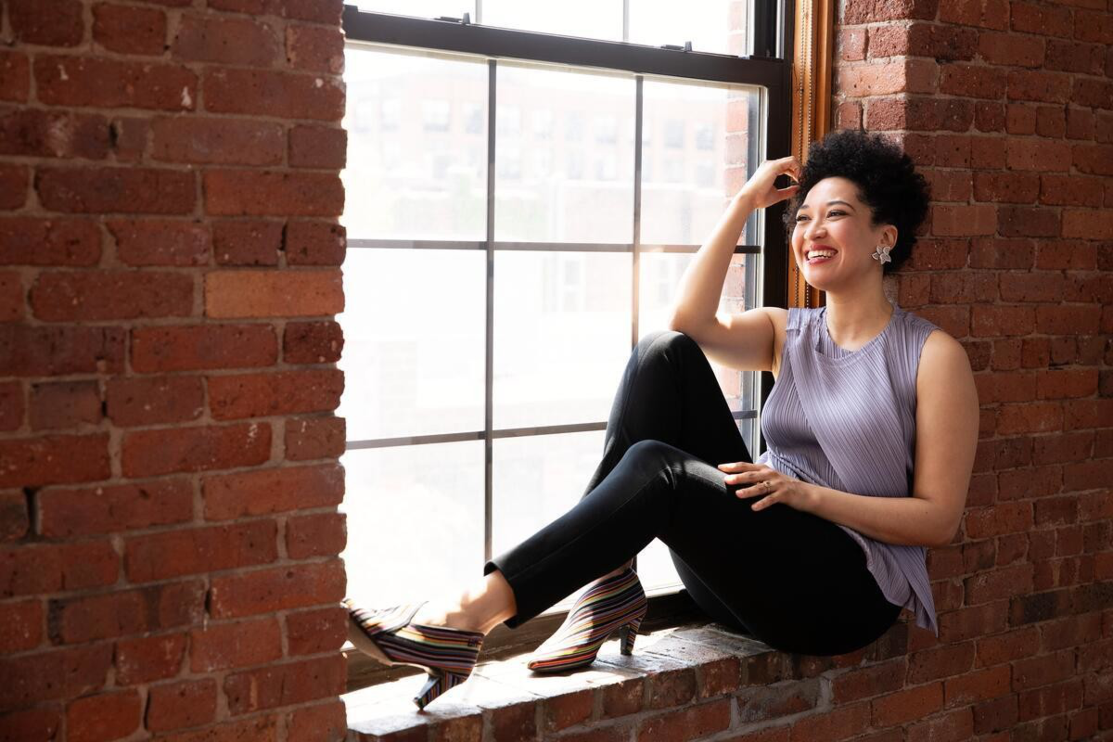 A smiling woman with curly hair sits on a brick windowsill, wearing a sleeveless purple top, black pants, and striped heeled shoes.