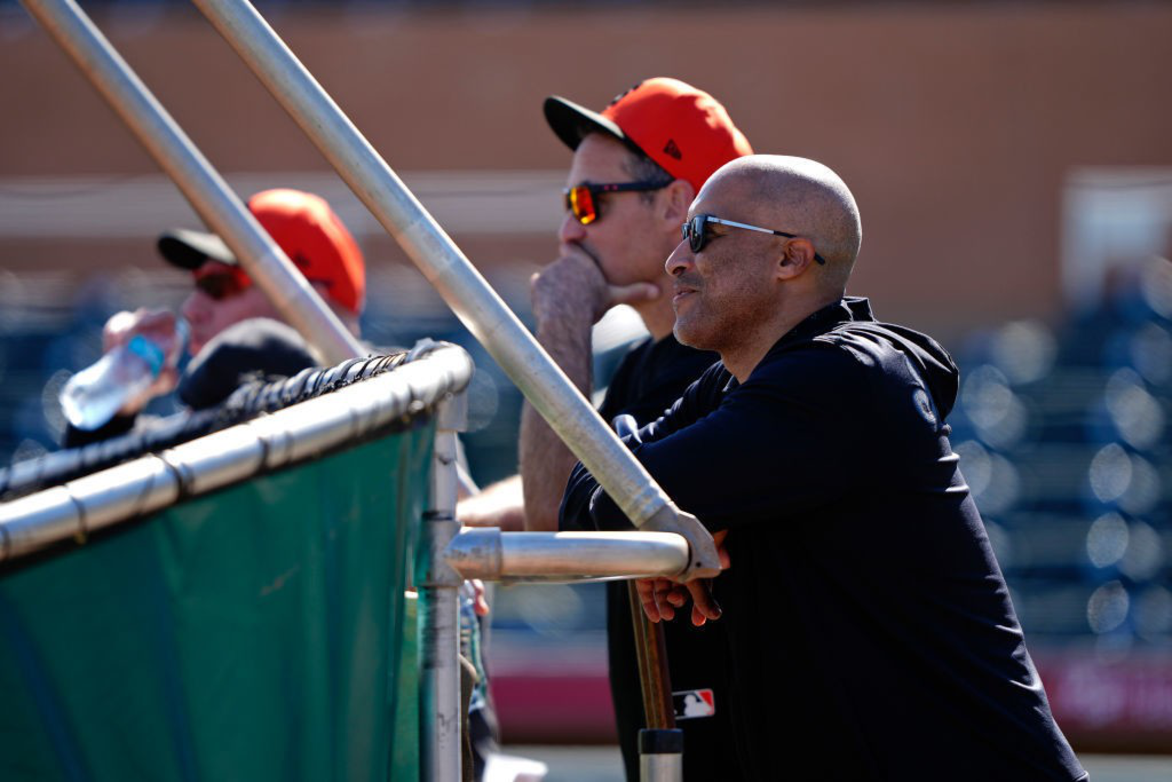 Three people stand near a railing at a baseball field, two wearing orange caps, one with sunglasses, looking focused. The sun casts a bright light on them.
