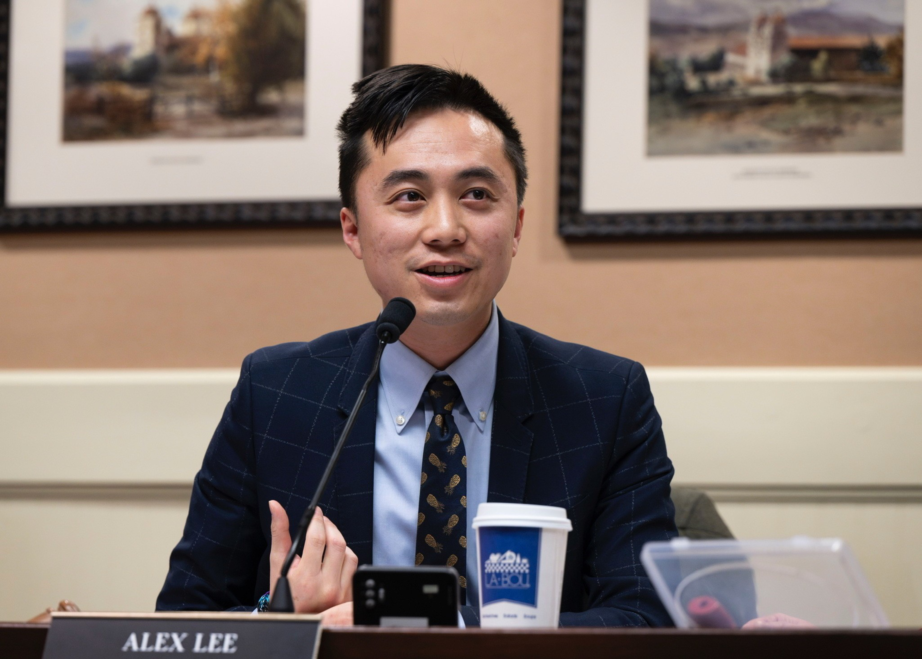 A man in a suit speaks at a desk with a microphone, nameplate, and coffee cup in front of framed art.