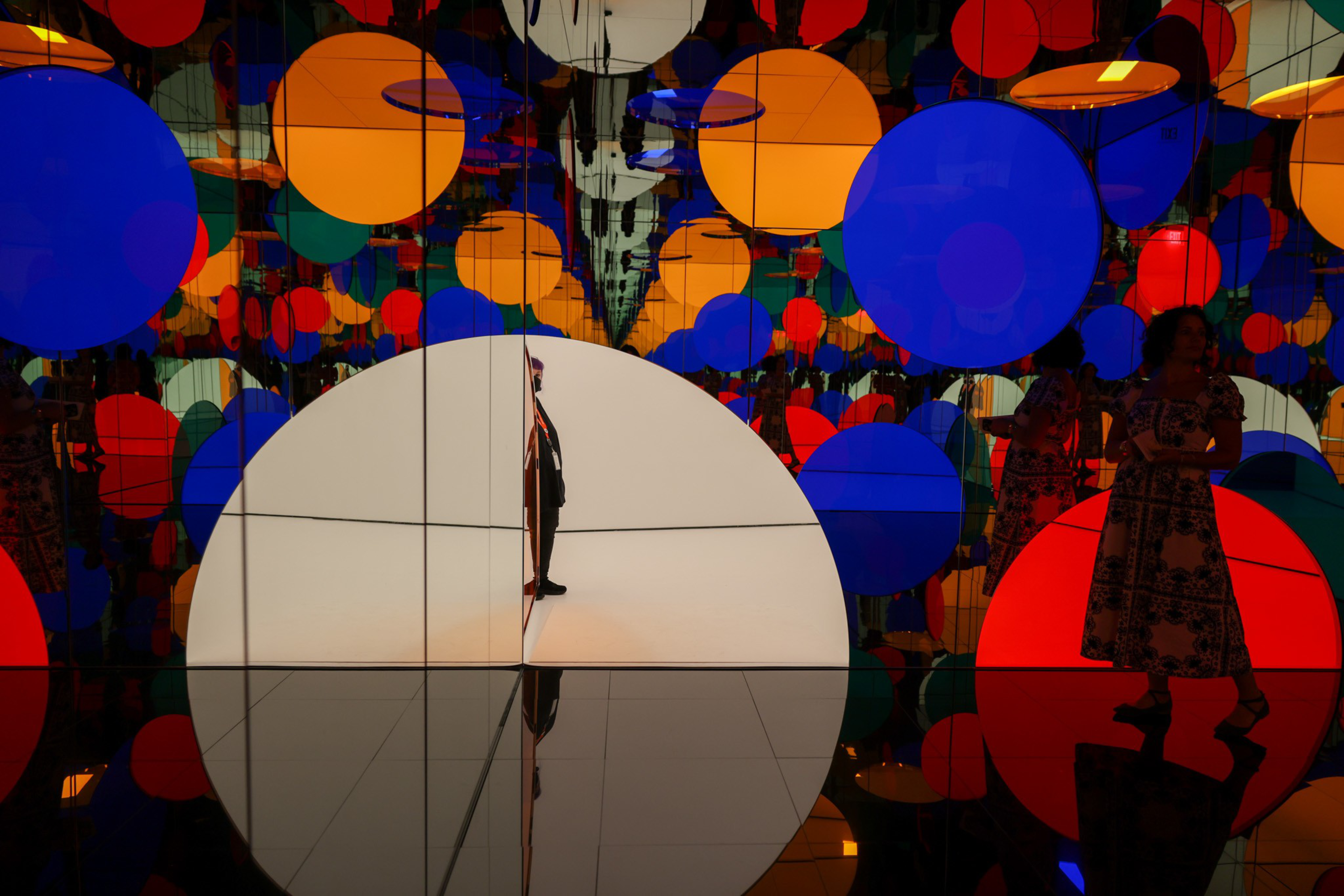 A room filled with large colorful circles in orange, blue, red, and white, reflecting in mirrors with two partially visible people standing inside.