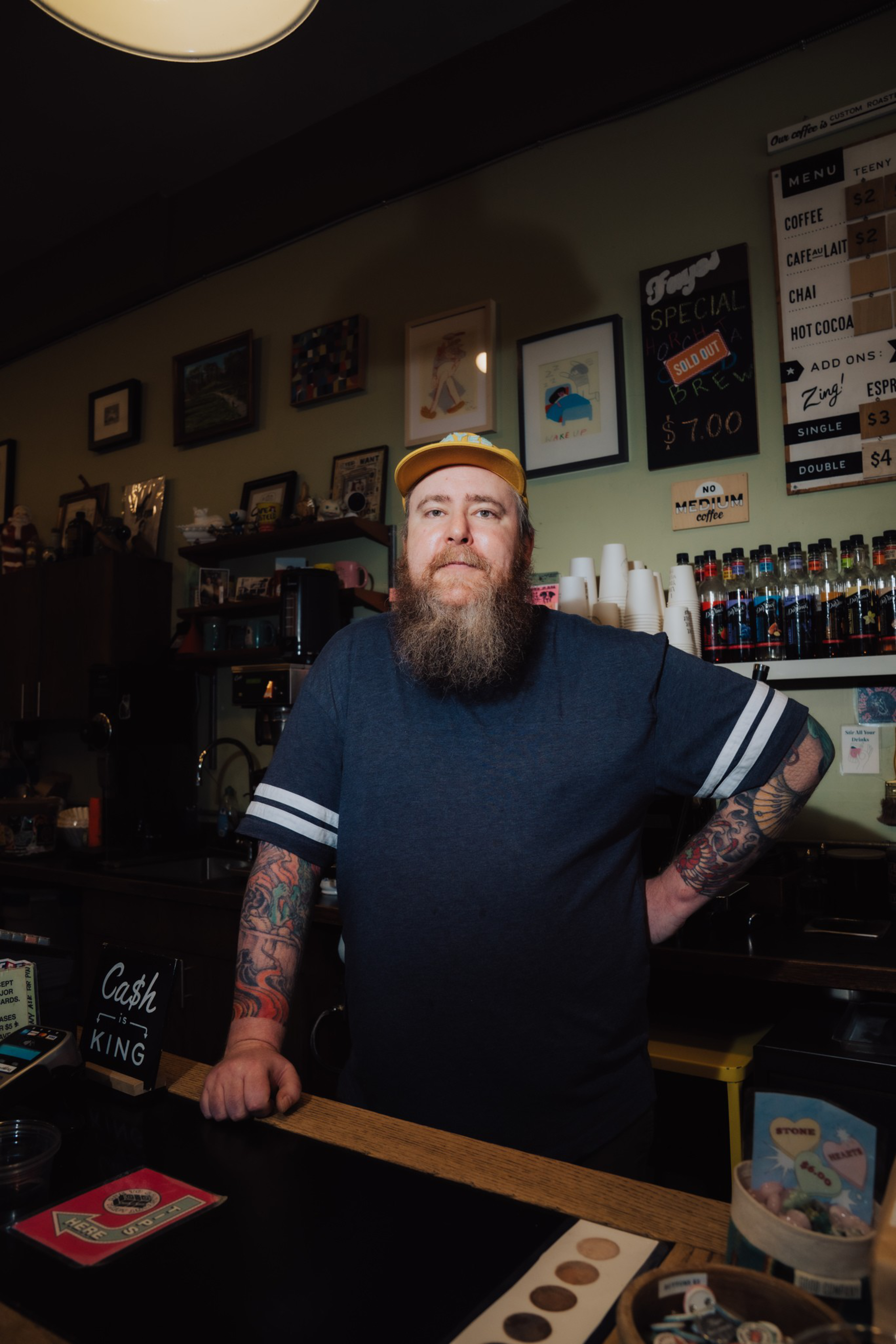 A bearded man with tattoos, wearing a yellow cap and navy shirt, stands behind a coffee shop counter with a menu and cups behind him.