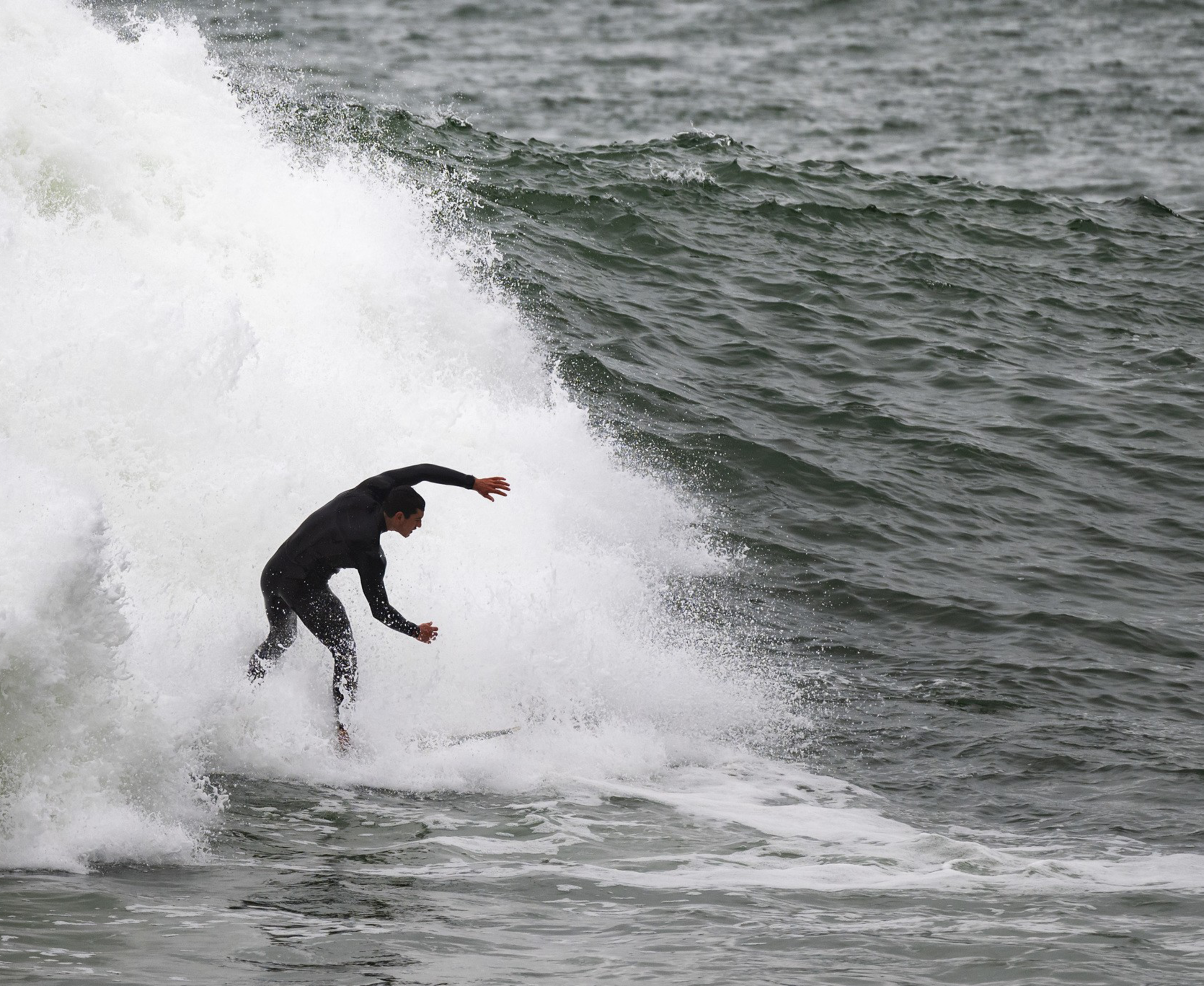 A surfer in a black wetsuit rides a powerful, frothy wave in the ocean, with water splashing dramatically around them.
