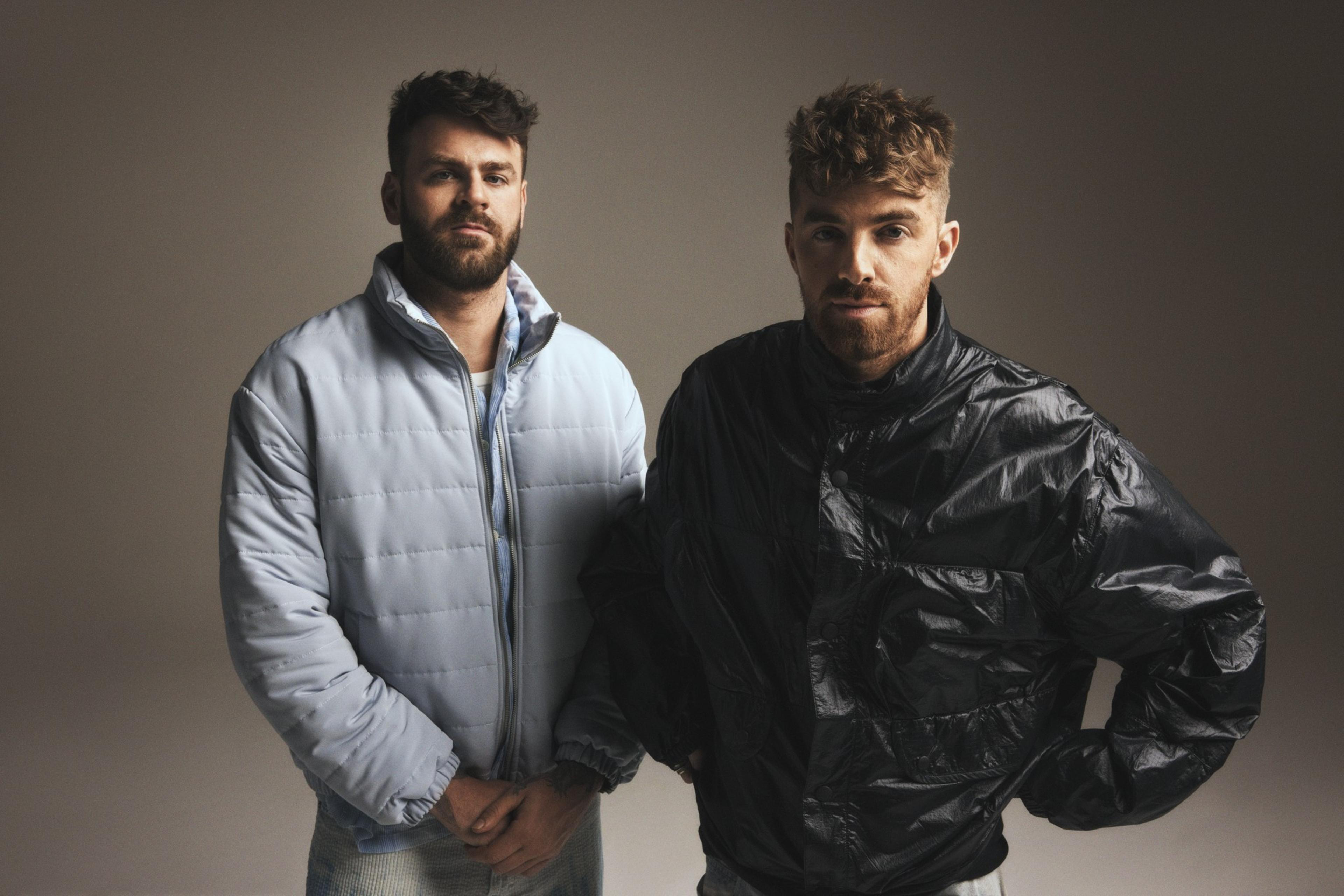 Two men stand side by side, one in a light blue puffer jacket and the other in a dark bomber jacket, both with short hair and beards against a plain backdrop.