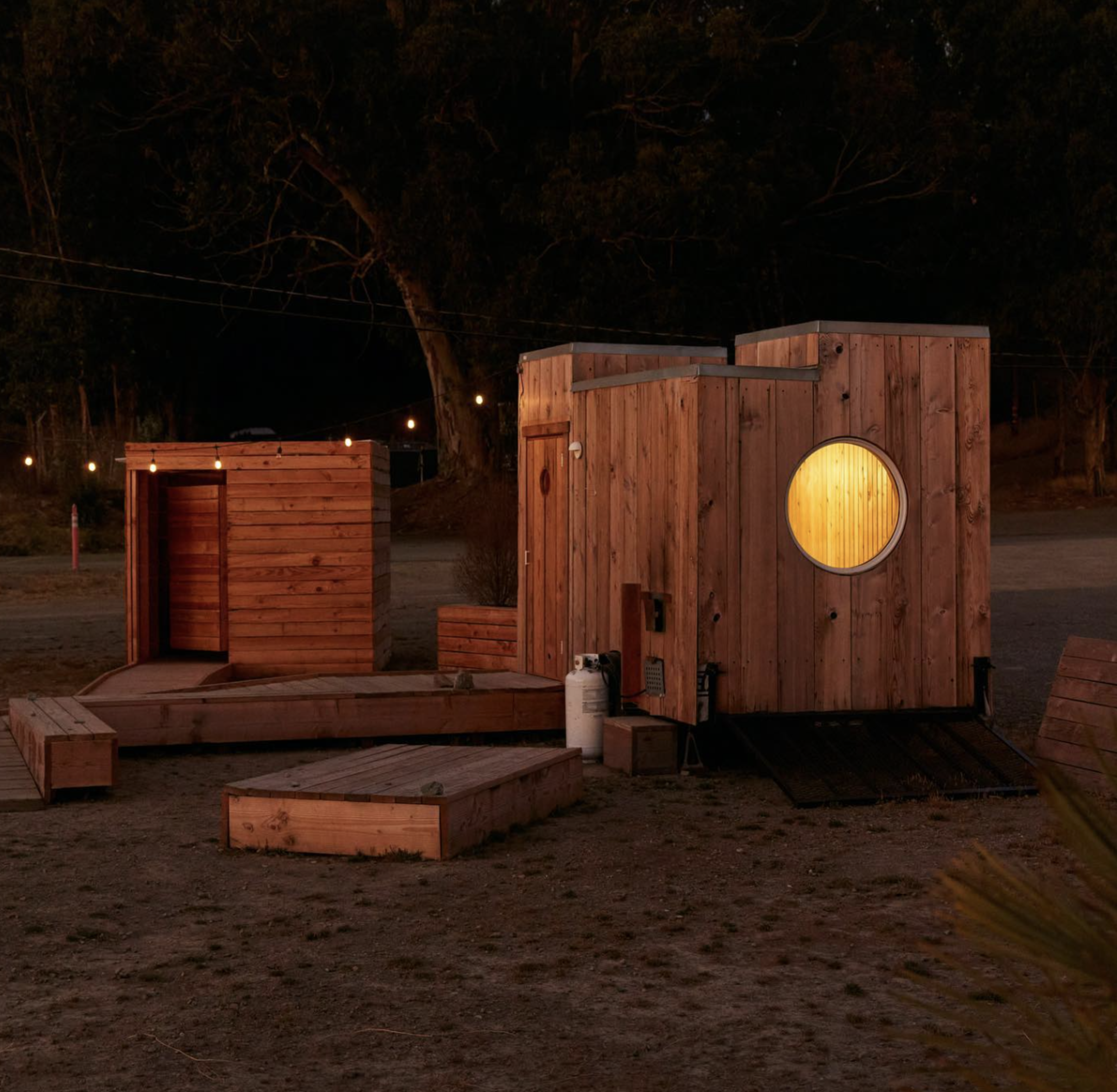 Two small wooden structures with warm lights and a circular window stand outdoors at night, surrounded by trees and strings of lights.