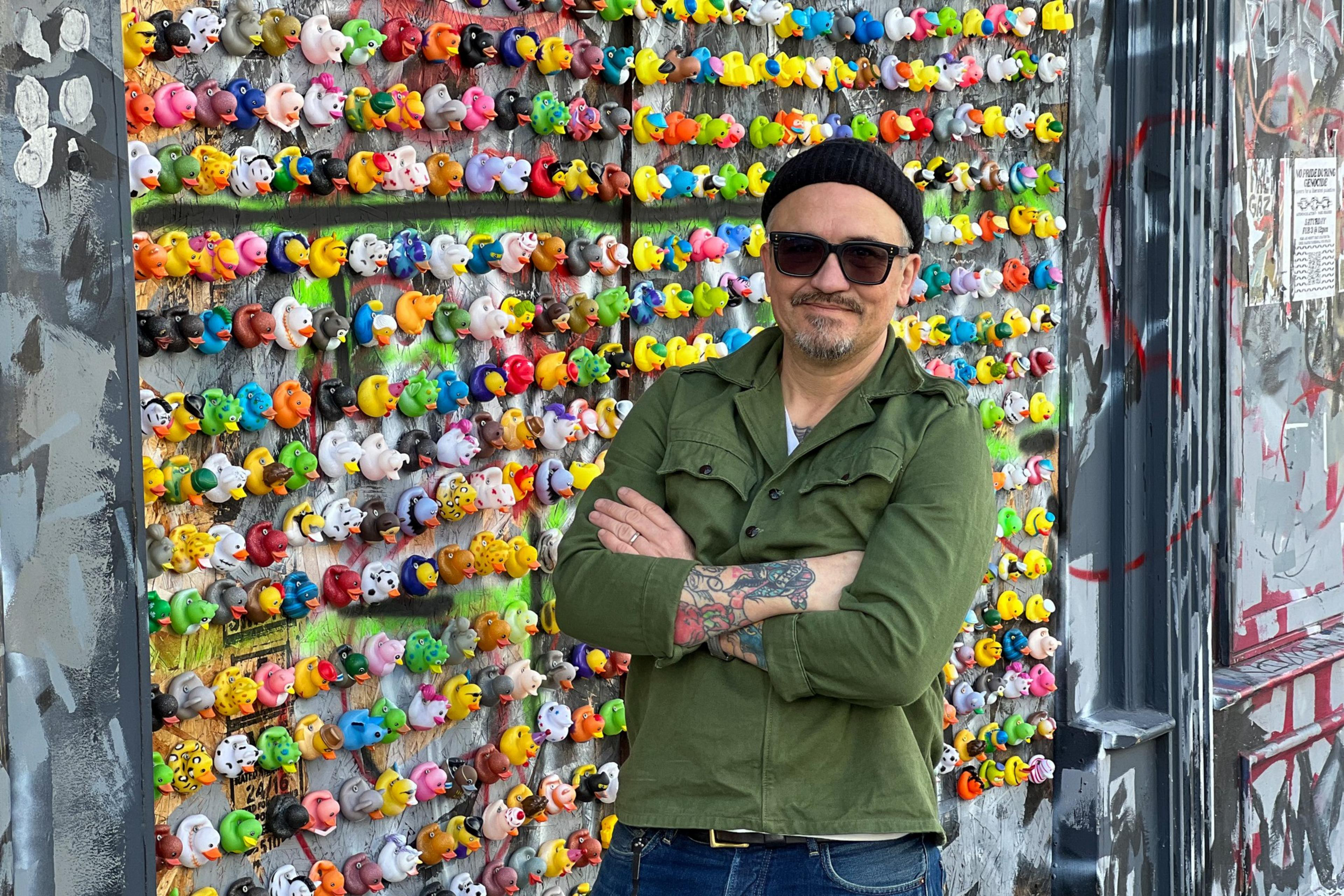 A man stands arms crossed before a vibrant wall of colorful rubber ducks.