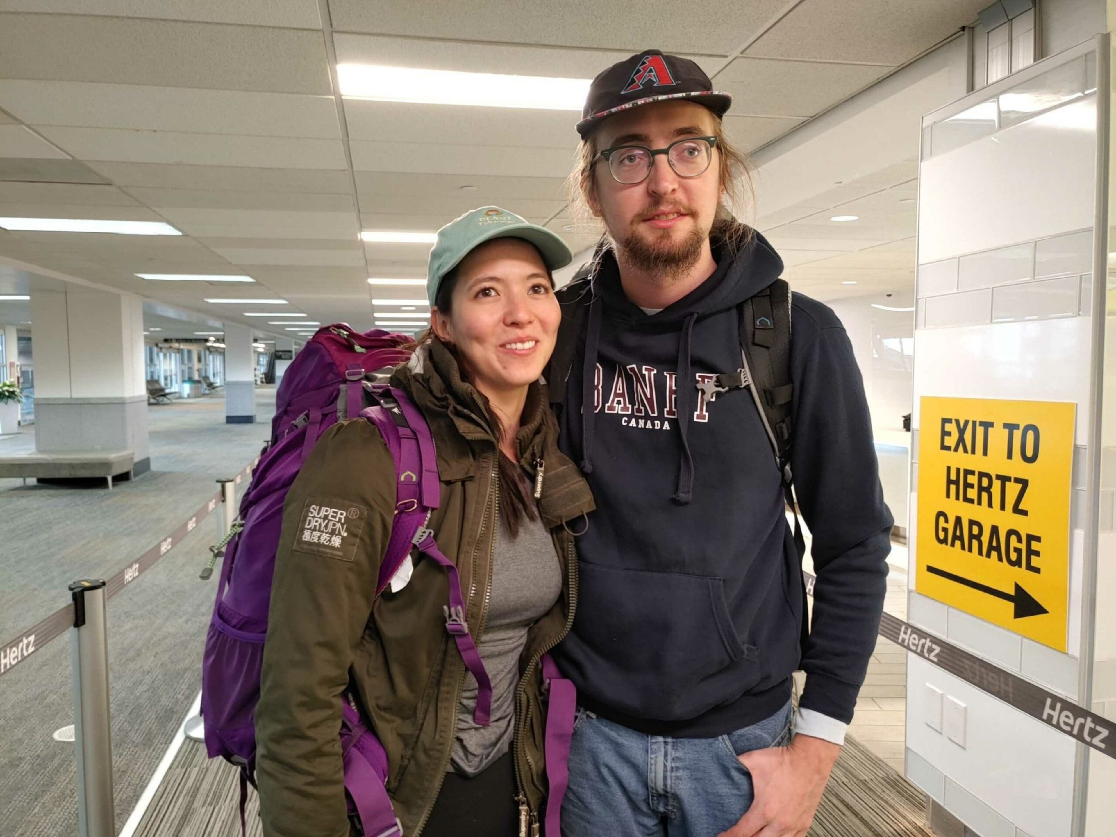 A smiling couple with backpacks stands close together in an airport, next to a yellow sign that says "Exit to Hertz Garage."