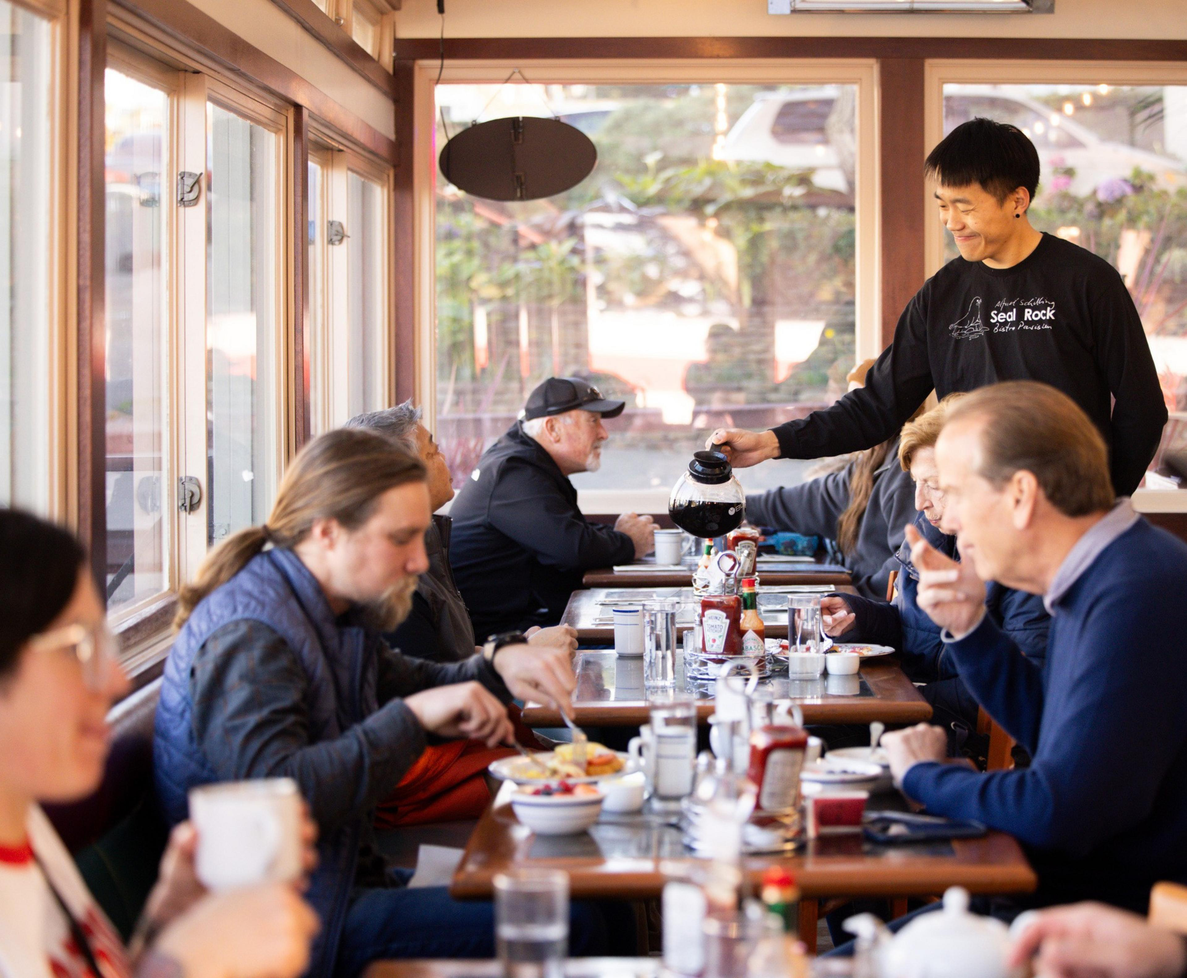 In a cozy diner, a waiter refills coffee for patrons sitting at wooden tables. The scene is lively, with people engaged in conversation and enjoying their meals.