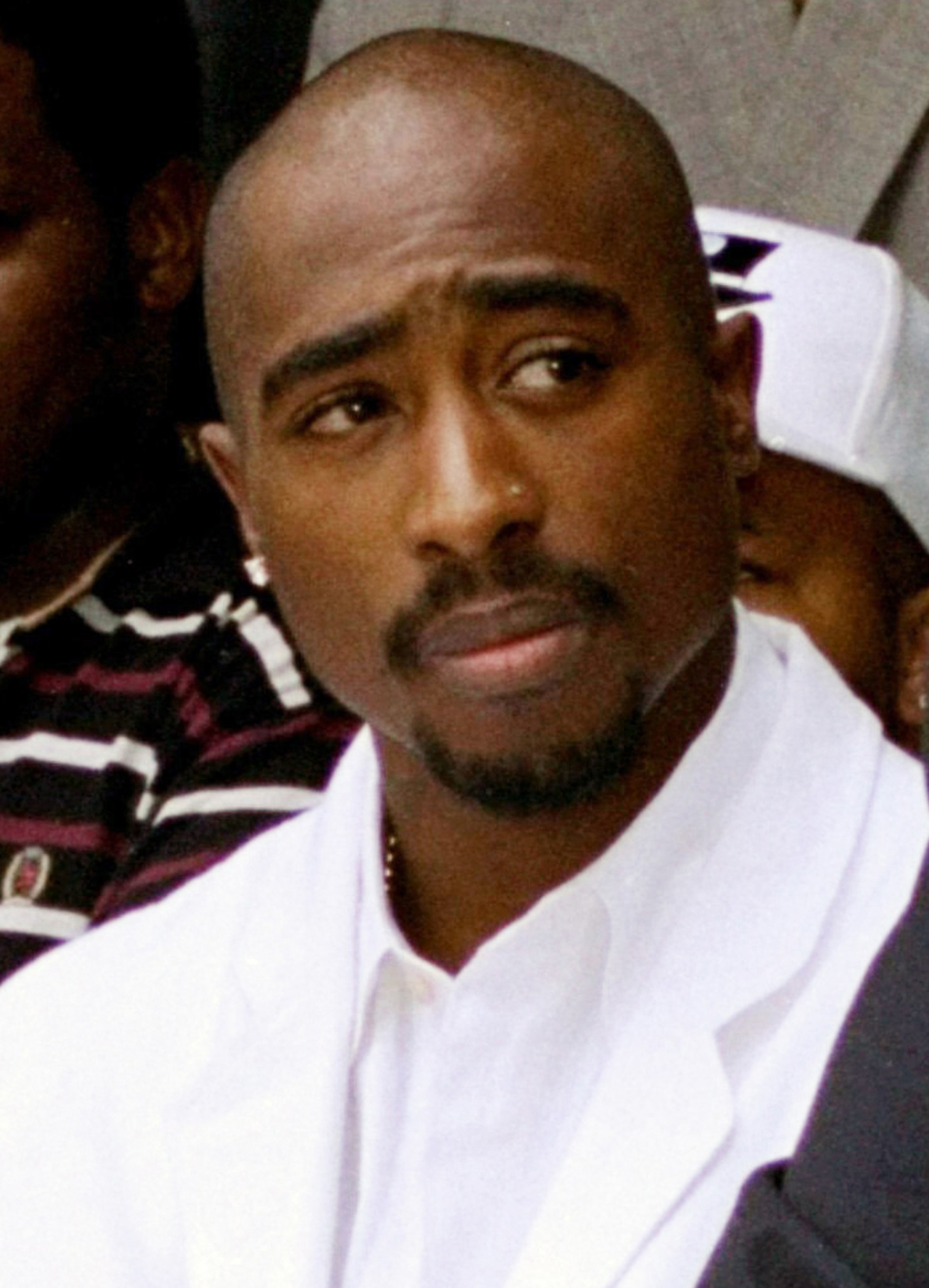 A man with a shaved head, mustache, and goatee wears a white jacket and white shirt, looking slightly to his left with a calm expression.
