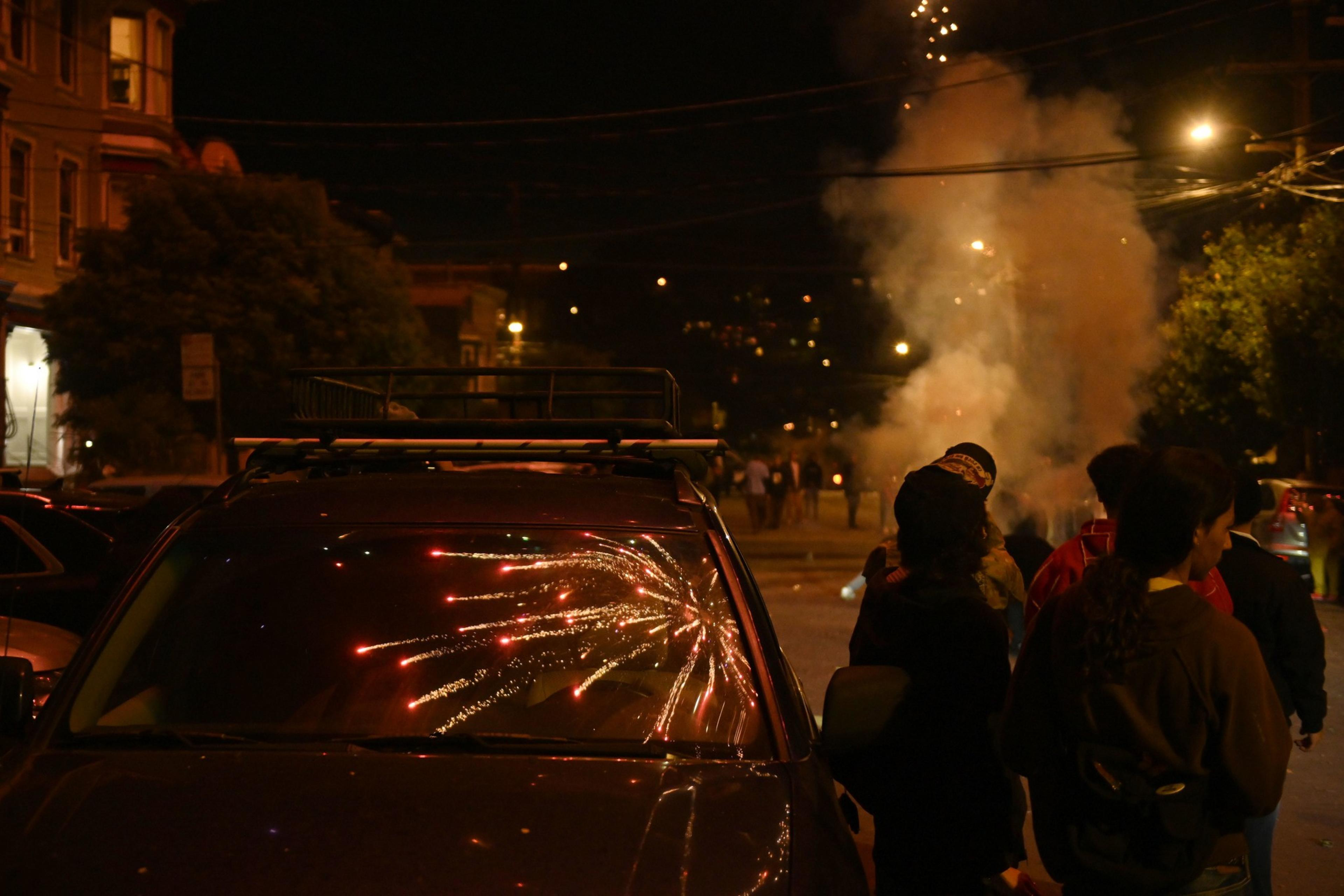 A person in a beanie and mask is lighting a firecracker, which is sparking, at night.