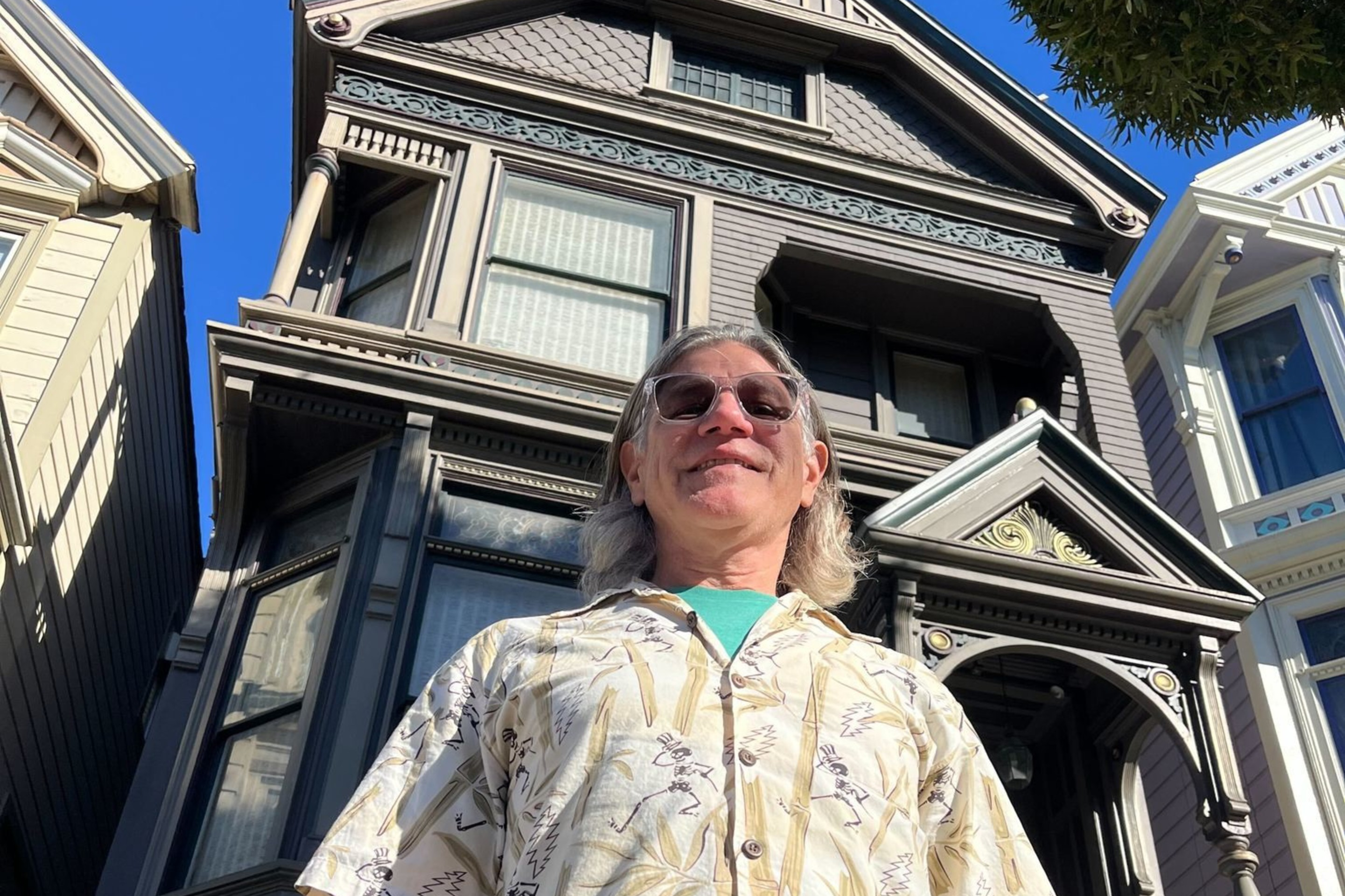 A person in a patterned shirt stands in front of a detailed, multi-story Victorian house with a bright blue sky above.