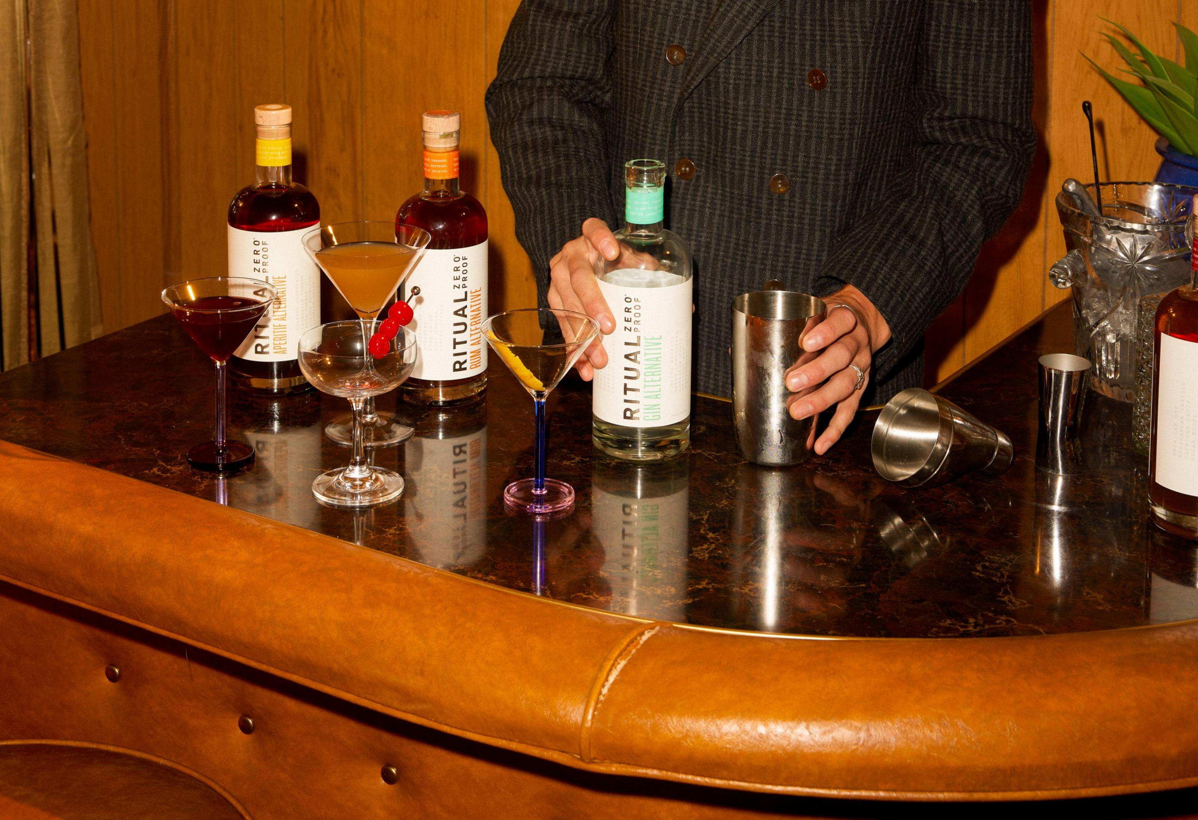 A person is behind a bar with various cocktail glasses and nonalcoholic spirits, preparing a drink with a shaker.