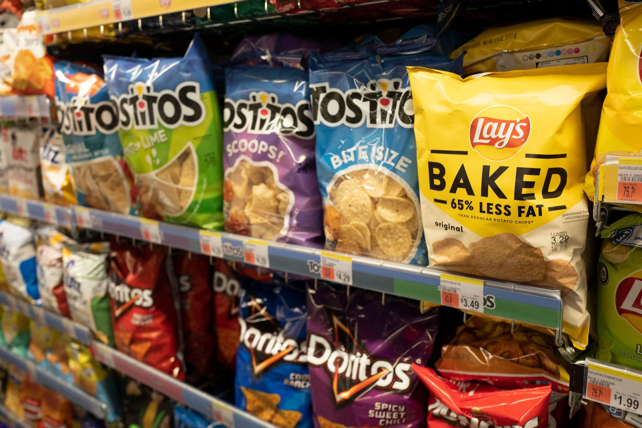 Rows of blue, green, red, and yellow bags of Tostitos, Lay's Baked, Doritos, and other chips displayed on store shelves.