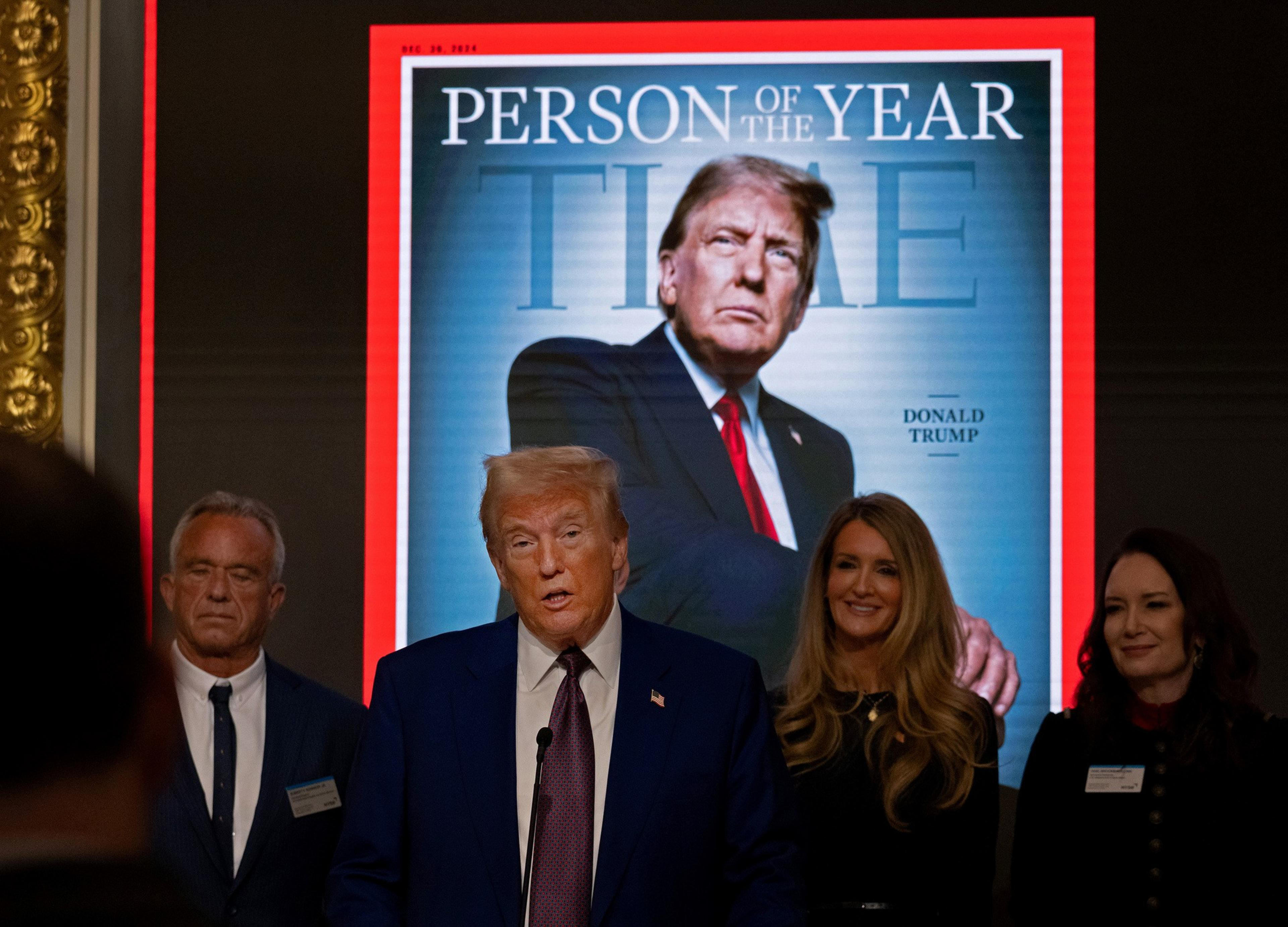 A man speaks at a podium, flanked by others, in front of a projected magazine cover featuring himself titled &quot;Person of the Year.&quot;