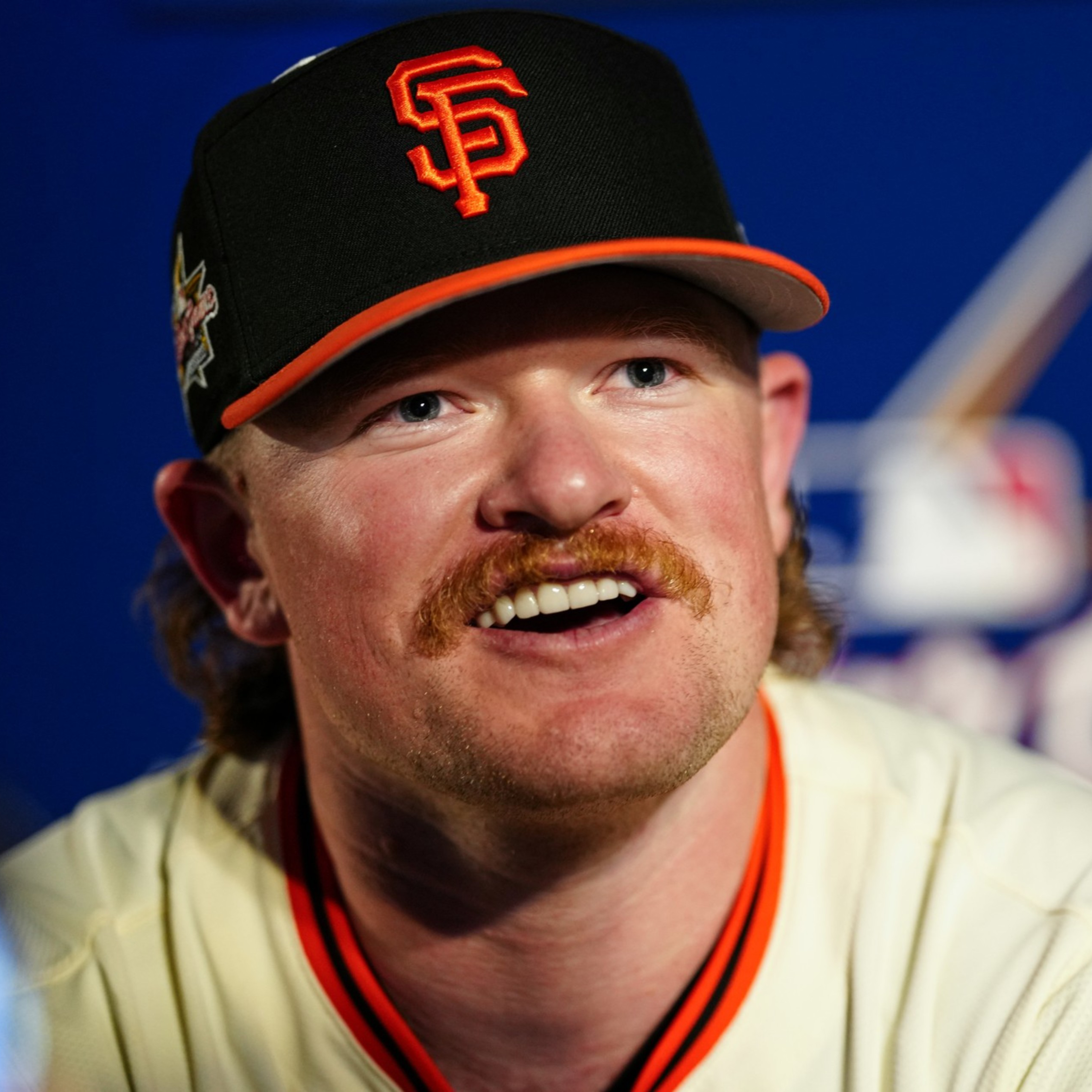 A man with a mustache wears a black SF cap and a baseball jersey. He is smiling and looking slightly upwards, with a blurred sporting event logo behind him.